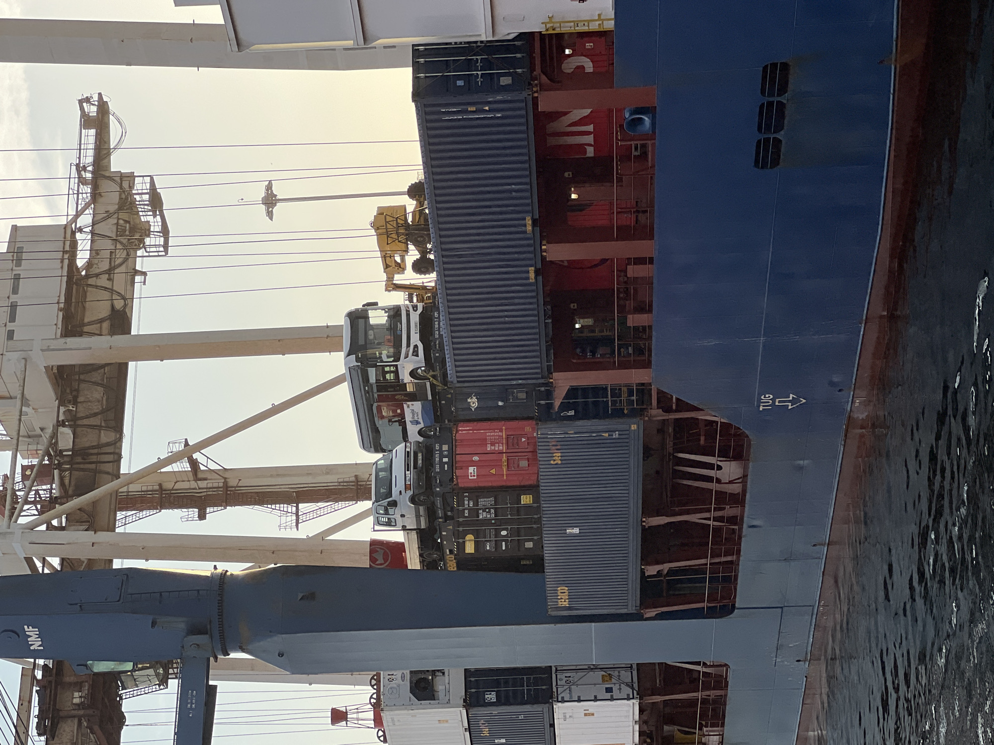 The image depicts a large cargo ship docked at the port of Matosinhos, Portugal. The ship is laden with shipping containers stacked high on its deck. On top of some of these containers, there are several vehicles, including a white truck with a flatbed and a white bus with "Hospital" written on its side. A yellow construction vehicle is also visible. The ship itself is blue with some red accents. In the background, massive port cranes are present, suggesting active cargo operations. The sky is overcast, and the water in the foreground shows gentle ripples. The scene captures a moment of logistical activity at a maritime port.