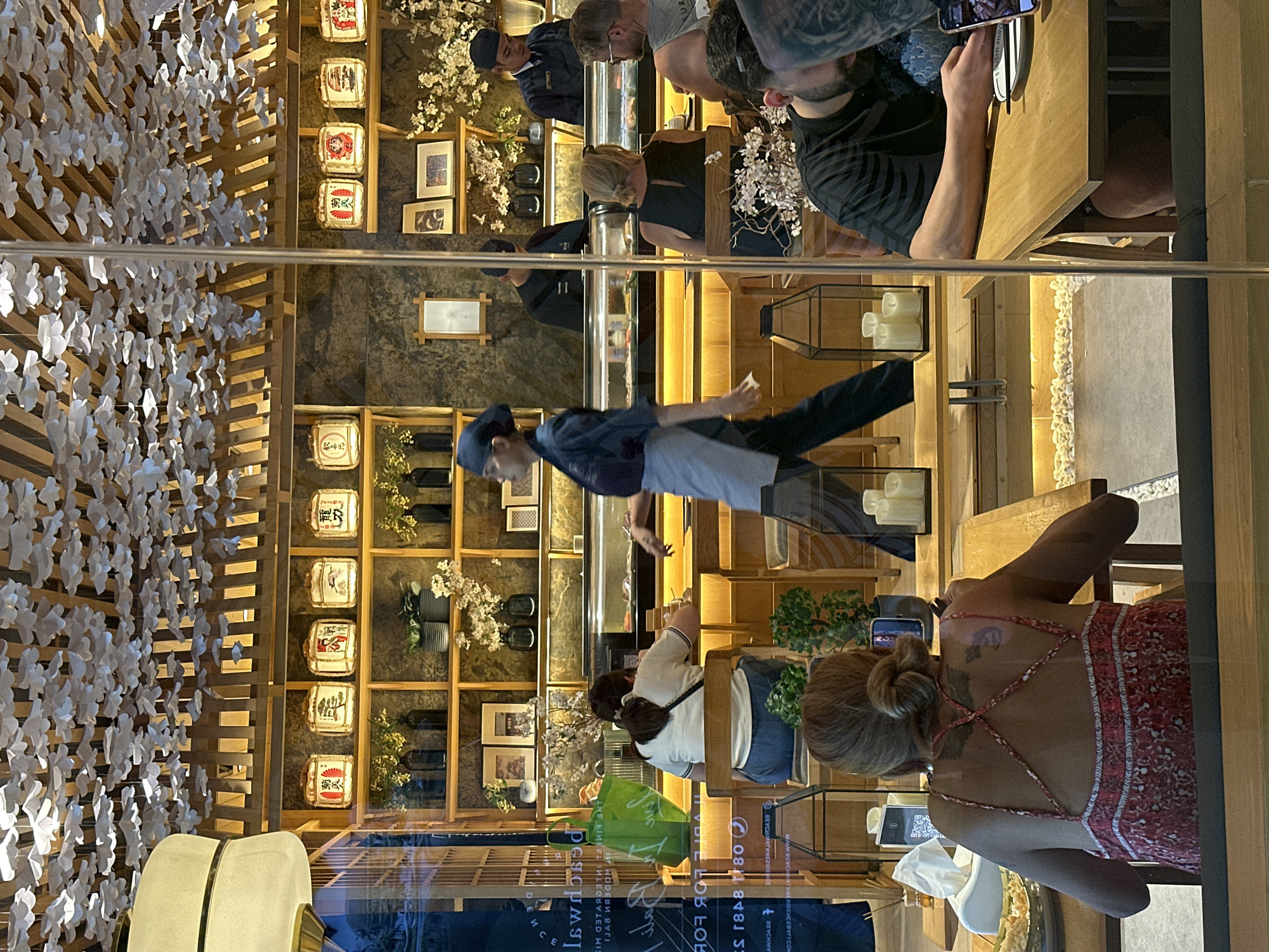 This photo was taken through the front window of a Japanese restaurant in Kuta, Indonesia. The view of the restaurant interior is reflected in the glass. The front window has printed business details on it.
Inside, a long counter staffed by Japanese chefs can be seen in the middle of the space. Behind the counter is shelving with several framed pictures, decorative sake barrels, and flower vases. White, origami-like bird decorations hang from the left side of the ceiling.
Several restaurant customers are seated at tables to the right and bottom right of the image. Some are actively looking at their phones. A woman is wearing a light red dress with spaghetti straps and has a tattoo on her back.