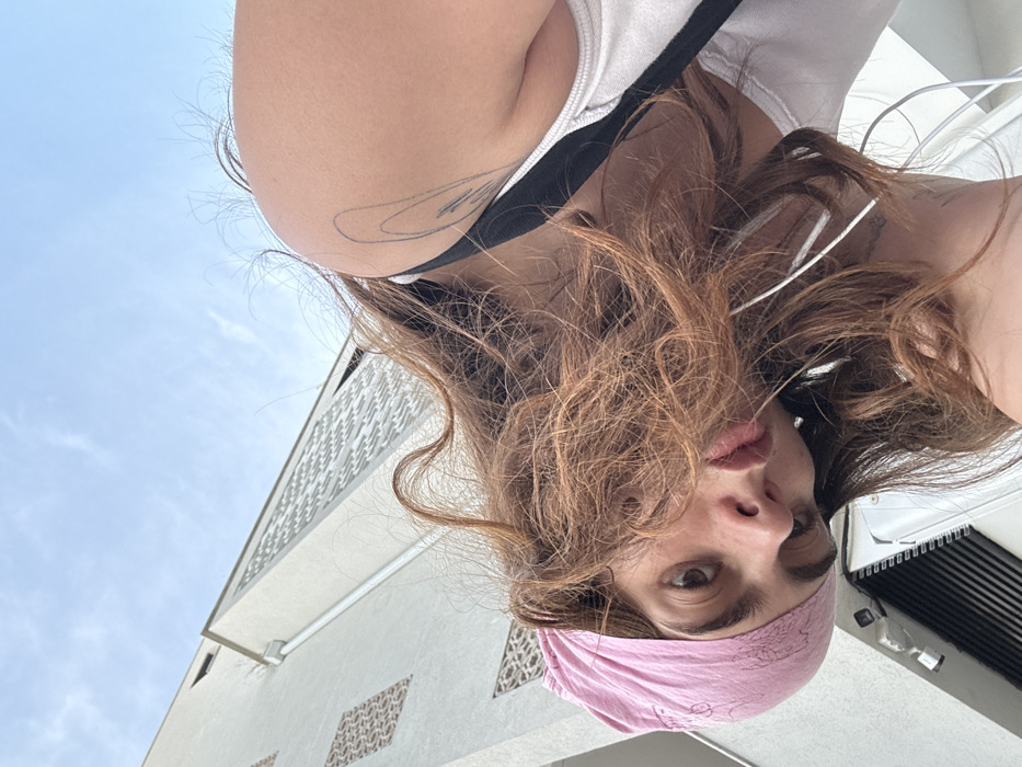 A low-angle, upward-facing image captures a young female subject in the foreground, with a building facade and sky in the background. The subject's head is tilted to the left, and her upper torso is partially visible. She has long, wavy, reddish-brown hair, with strands obscuring parts of her face. A pink bandana, featuring a faint patterned design, is worn on her head. Her dark eyes are directed towards the camera, and her lips are slightly parted. White wired earphones are visible, extending from her ears downwards. She wears a white tank top with a black strap visible over her right shoulder. A black outline tattoo, composed of multiple curved lines, is present on her upper right arm.
The background features a light-colored, multi-story building, consistent with modern residential or commercial architecture found in Tel-Aviv, Israel. The building's facade displays two distinct decorative patterns: a darker geometric pattern on a lower section and a lighter, intricate, perforated screen-like pattern on a higher section. A white gutter runs vertically along the building's side. The upper right portion of the image shows a clear blue sky with minimal scattered white clouds. The exact location within Tel-Aviv is not identifiable from the visible architectural details.