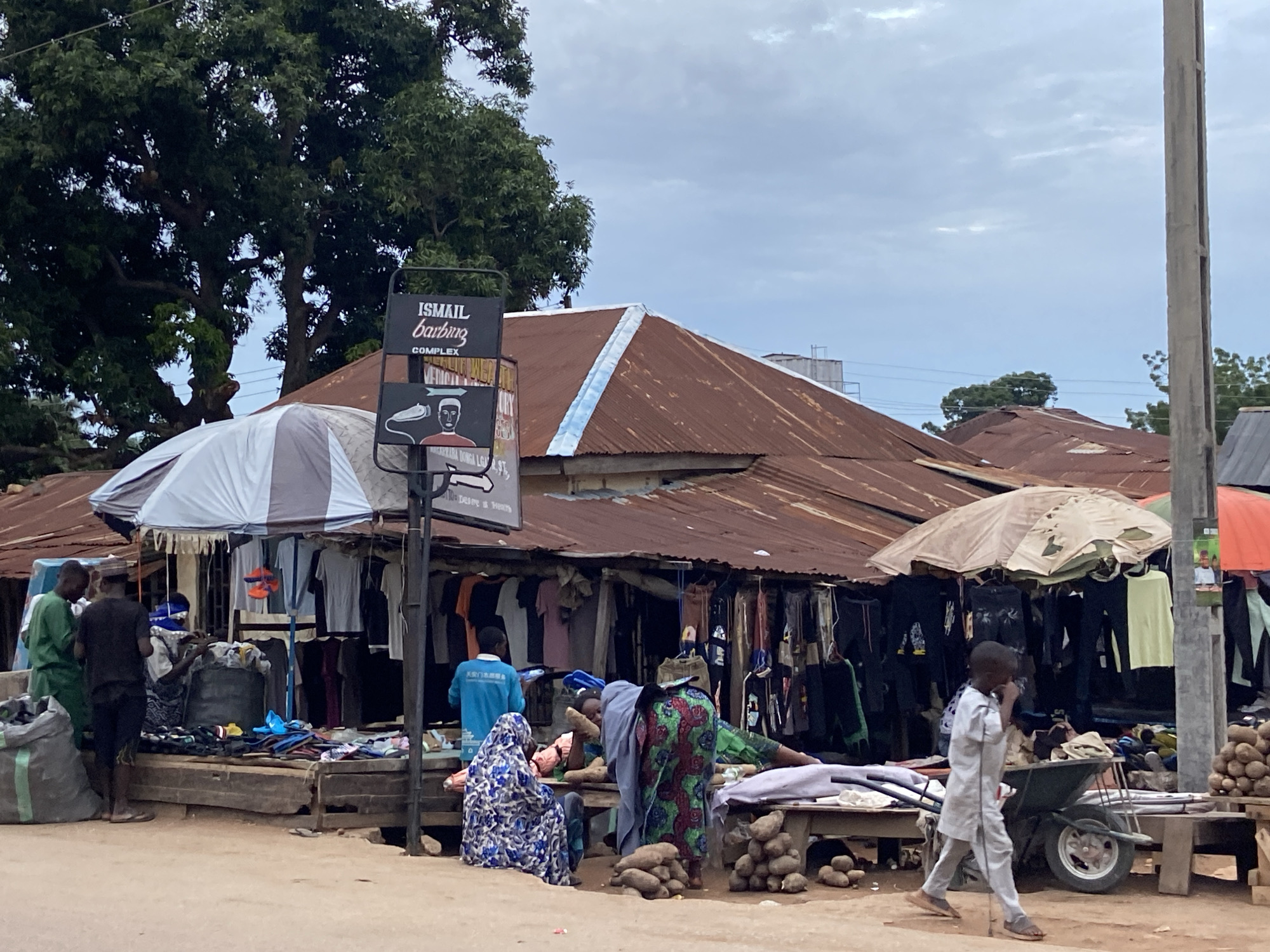 The image depicts a bustling street scene in Mararaba, Nigeria. The foreground shows a dirt road with several people engaged in commerce. A woman in a blue and white hijab sits on a low wooden bench near a pile of yams. Another woman stands nearby, wearing a colorful, patterned dress. To the left, a man in green stands near a vendor displaying clothes and shoes. A person in white walks past the scene to the right, pushing a wheelbarrow.
Behind them, a series of open-air shops are visible, each displaying various items, including clothing. Umbrellas provide shade over some of the stalls. The shops are housed under a corrugated metal roof that is faded with rust. A sign for "ISMAIL barbering COMPLEX" is visible. A large tree is located on the left side of the image. Utility poles are visible in the background. The sky is overcast.