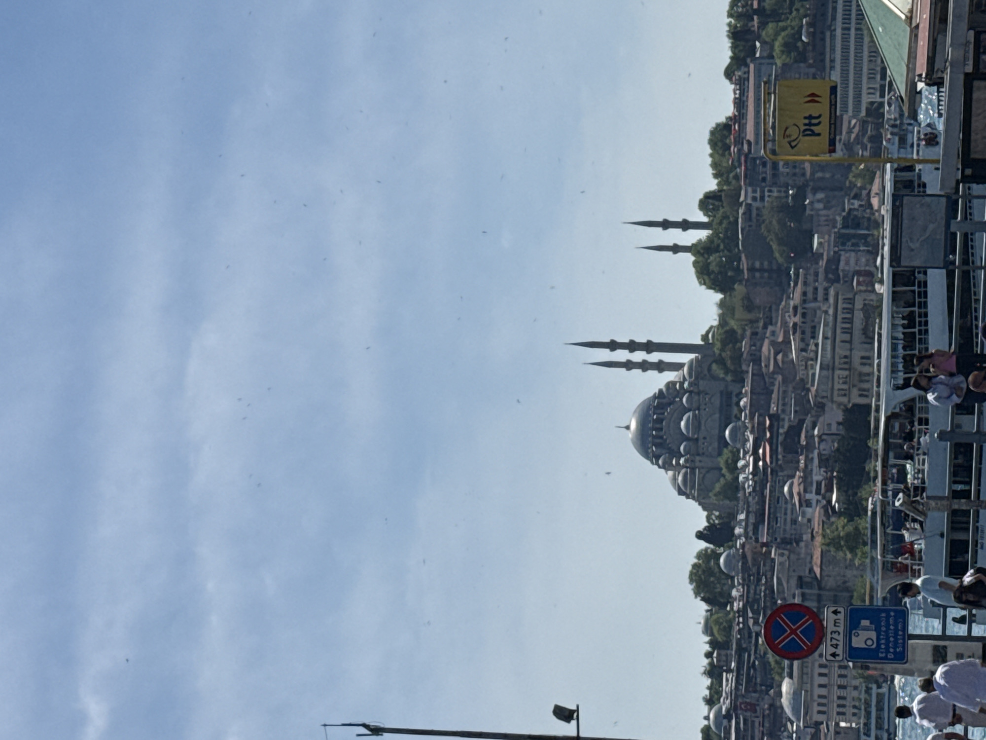 The image depicts a cityscape featuring a prominent mosque with multiple domes and tall minarets. In the foreground, a bridge or walkway is visible, lined with metal railings. People are gathered on the bridge, some appearing to be observing the view. Road signs are also present, including a "no stopping" sign and another with Turkish script, indicating an electronic surveillance system. The sky is blue with scattered clouds. A yellow Ptt sign is seen in the midground. The location is likely Istanbul, Turkey, given the architectural style of the mosque, which resembles the Süleymaniye Mosque.