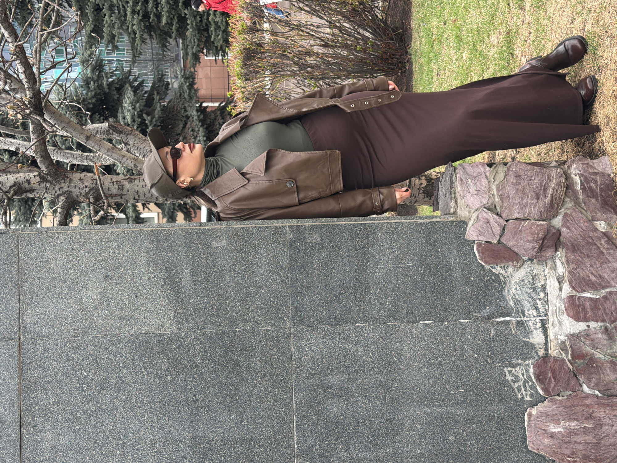 A woman is leaning against a large stone structure in Krasnoyarsk, Russia. She wears a brown leather jacket, a green turtleneck, a brown skirt, a cap, and sunglasses. The structure consists of large, gray granite slabs on the left and a decorative arrangement of red and white rocks on the right. Behind the woman, there is a grassy area with trees and bushes. Other people are in the background.