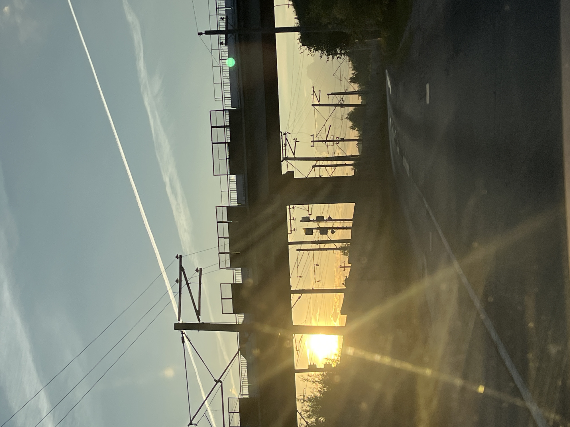 The image depicts a road with a railway overpass in Vasilyevo, Russia, during sunset. The sun is low on the horizon, casting a warm glow and creating lens flares that streak across the scene. The overpass is supported by concrete pillars and has a walkway with railings. Overhead, there are numerous power lines and catenary wires for the electric train system. In the sky, contrails from an aircraft are visible against a pale blue and yellow gradient. The road surface is asphalt, with white lane markings. There are trees and vegetation on either side of the road and beneath the overpass. The overall impression is one of a transitional space where infrastructure meets nature at dusk.