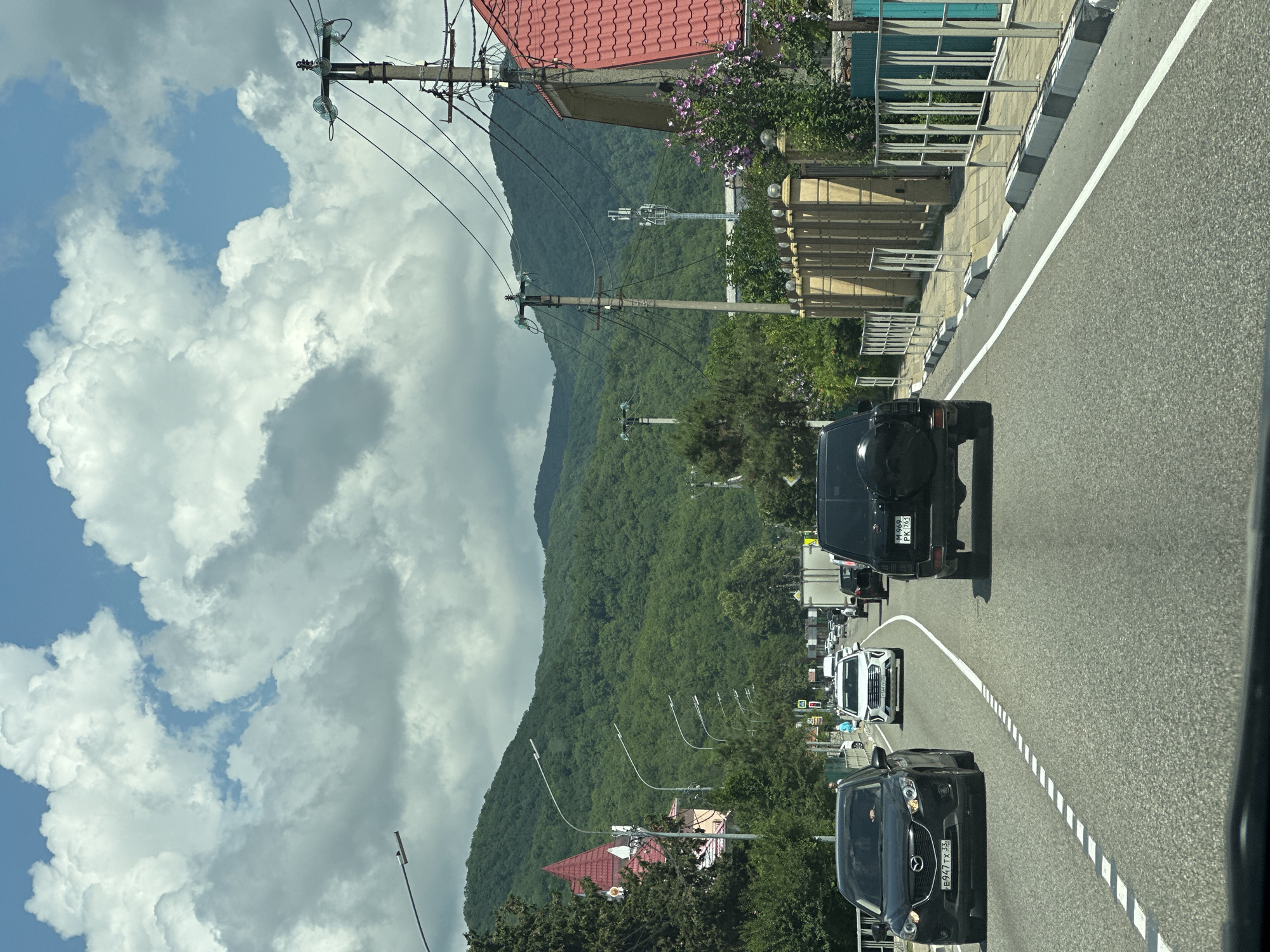 A rear-view perspective from a vehicle on a paved road in Shepsi, Russia, shows a traffic scene under a partly cloudy sky with large, fluffy white clouds against a blue sky. The road has two lanes in each direction, separated by a dashed white line. Several vehicles are visible, including a black SUV in the foreground, a black Mazda CX-5 with a license plate E947TX 33 on the left lane, and a white sedan ahead with a license plate E564XY 93. In the distance, mountains covered in dense green forest form the backdrop. Along the right side of the road, there is a sidewalk with a light brown fence, ornamental lamps, and a building with a red tiled roof. Utility poles with wires and what appears to be a cell tower are also visible. On the left side of the road, a building with a distinctive red pointed roof is present, along with more utility poles and trees. The scene suggests a moderately busy traffic day in a semi-urban or suburban area with significant natural scenery.