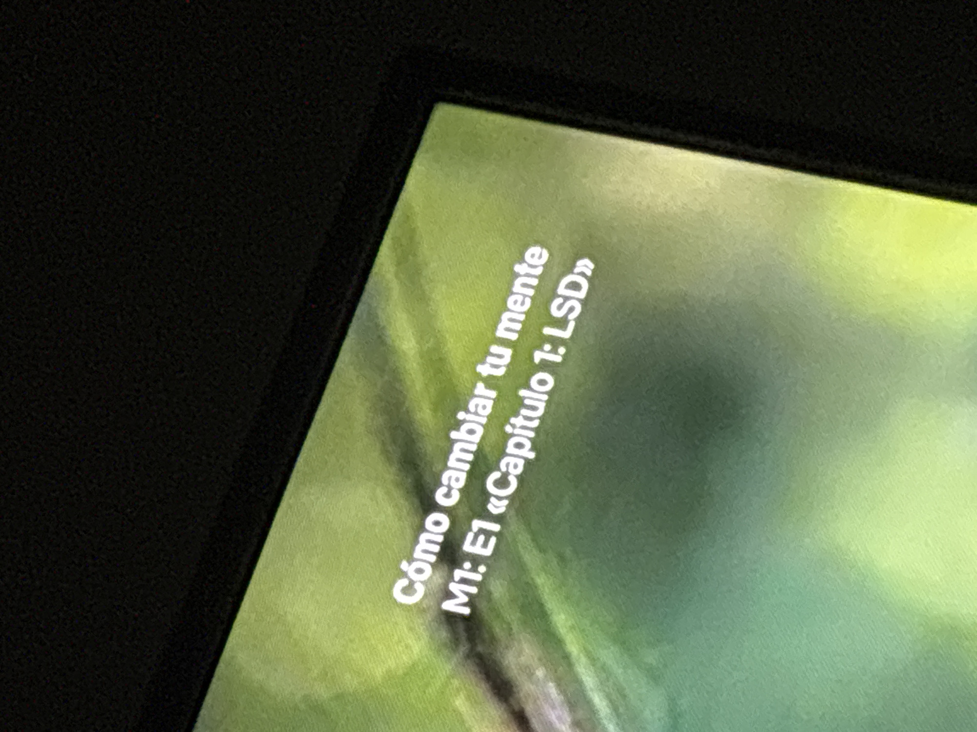 The image shows a close-up of a screen displaying text in Spanish: "Cómo cambiar tu mente M1: E1 «Capítulo 1: LSD»". The background of the screen appears to be a blurred image with green hues. The surrounding area is dark and out of focus. There is no indication of the location in the city of Buenos Aires, Argentina.