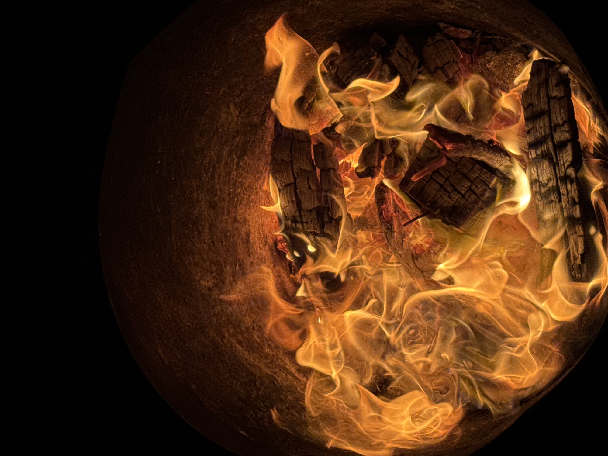 A close-up, high-angle shot captures the intense heat and flickering flames of a bonfire. The fire burns within a dark, round container, possibly a fire pit or cauldron. The primary subjects are the glowing embers, charred wooden logs, and the dynamic, orange and yellow flames that twist and writhe upwards. The logs are dark and deeply grooved, showing signs of intense heat and burning. Wisps of smoke are not prominently visible, but the swirling flames suggest a strong draft.
The scene is dark, with the exception of the fire itself, indicating it is likely nighttime or a very dimly lit environment. The background is predominantly black, creating a stark contrast that highlights the fiery activity. The texture of the container's interior is rough and uneven, suggesting a natural material like stone or thick metal.
There is no indication of people or specific weather conditions. The location context of Chaikovsky, Russia, does not provide specific visual cues within the image itself. The dominant emotions evoked by the image are warmth, intensity, and a sense of primal energy. The image focuses entirely on the visual spectacle of the burning fire, devoid of any textual elements.