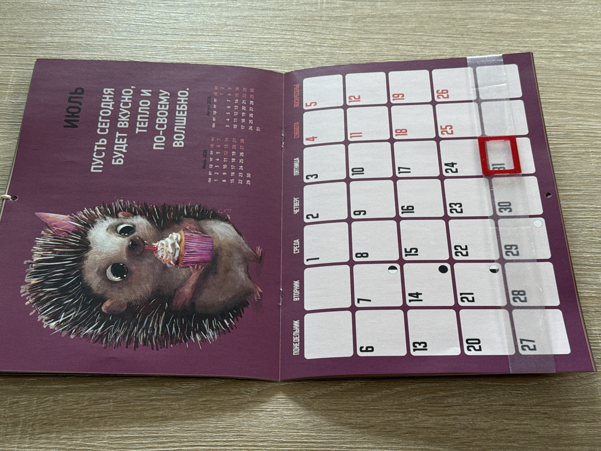 The image shows a wall calendar for July 2025, featuring a drawing of a hedgehog holding a cupcake. The calendar is hanging on a wooden wall. The top part of the calendar has the Russian word "ИЮЛЬ" (July) and a quote that reads "ПУСТЬ СЕГОДНЯ БУДЕТ ВКУСНО, ТЕПЛО И ПО-СВОЕМУ ВОЛШЕБНО." (Let today be delicious, warm, and magically unique). Below the illustration is a grid calendar for July 2025, with days of the week labeled in Russian (ПН, ВТ, СР, ЧТ, ПТ, СБ, ВС). The dates are displayed within individual boxes. The 31st of July is highlighted with a red square. There are also smaller calendars for June and August 2025 at the top. The calendar is attached to the wall with a metal clip at the top. The environment appears to be indoors, likely a home or office, given the wooden wall texture and the nature of the object. There are no people or specific landmarks visible that would indicate a location in Lyubertsy, Russia.