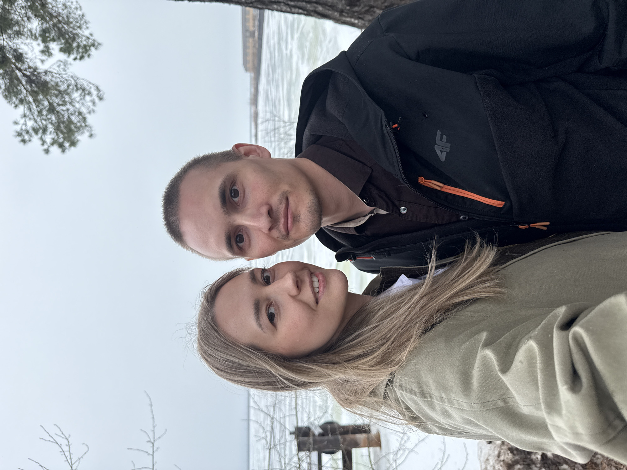 A selfie was taken by a man and a woman. The location appears to be outdoors, next to a body of water covered in ice. There is a sparse tree in the background and a small manmade structure next to the lake. Based on the location listed, it's possible this photo was taken in the city of Chaikovsky, Russia.
The man has short hair, and is wearing a brown button-up shirt under a dark jacket with a logo. The woman has blonde hair and is wearing a khaki colored jacket. Both are facing the camera.