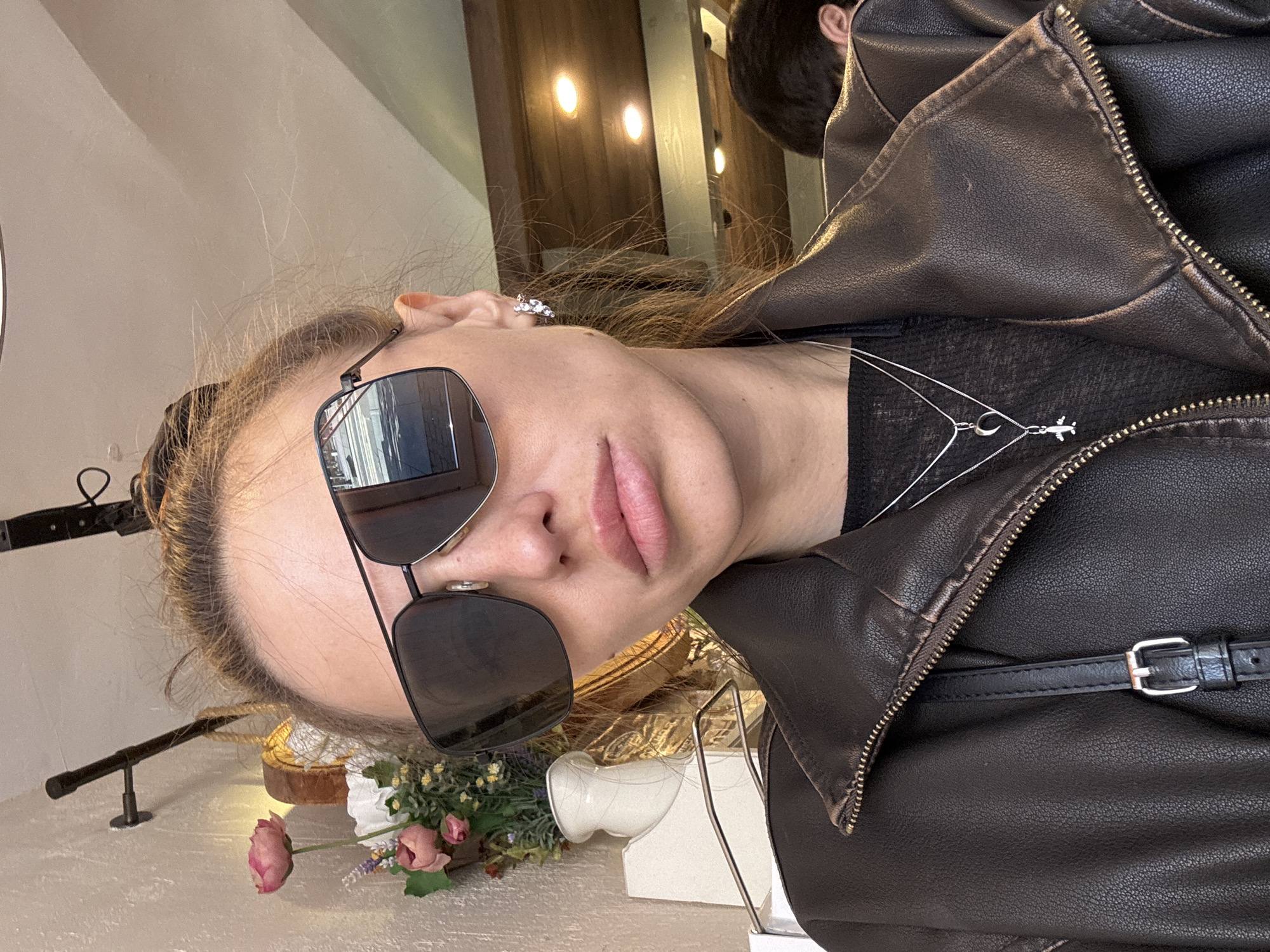 Here is a detailed description of the image:
**People:** The image primarily features a woman with fair skin, light brown hair, and sunglasses. A partial view of another person's head is visible behind her.
**Subjects/Objects:** The woman is wearing a black leather jacket and a black top with a silver necklace. She is wearing sunglasses with a black frame and reflective lenses. There is a small arrangement of flowers with pink roses on a light-colored surface. There's a display with lights, possibly in a shop.
**Actions/Interactions:** The woman is looking directly at the camera.
**Environment:** The setting appears to be indoors, potentially in a boutique or store in Saint Petersburg, Russia, given the style of the decor and lights, and based on the metadata, if available. The background is somewhat blurred.