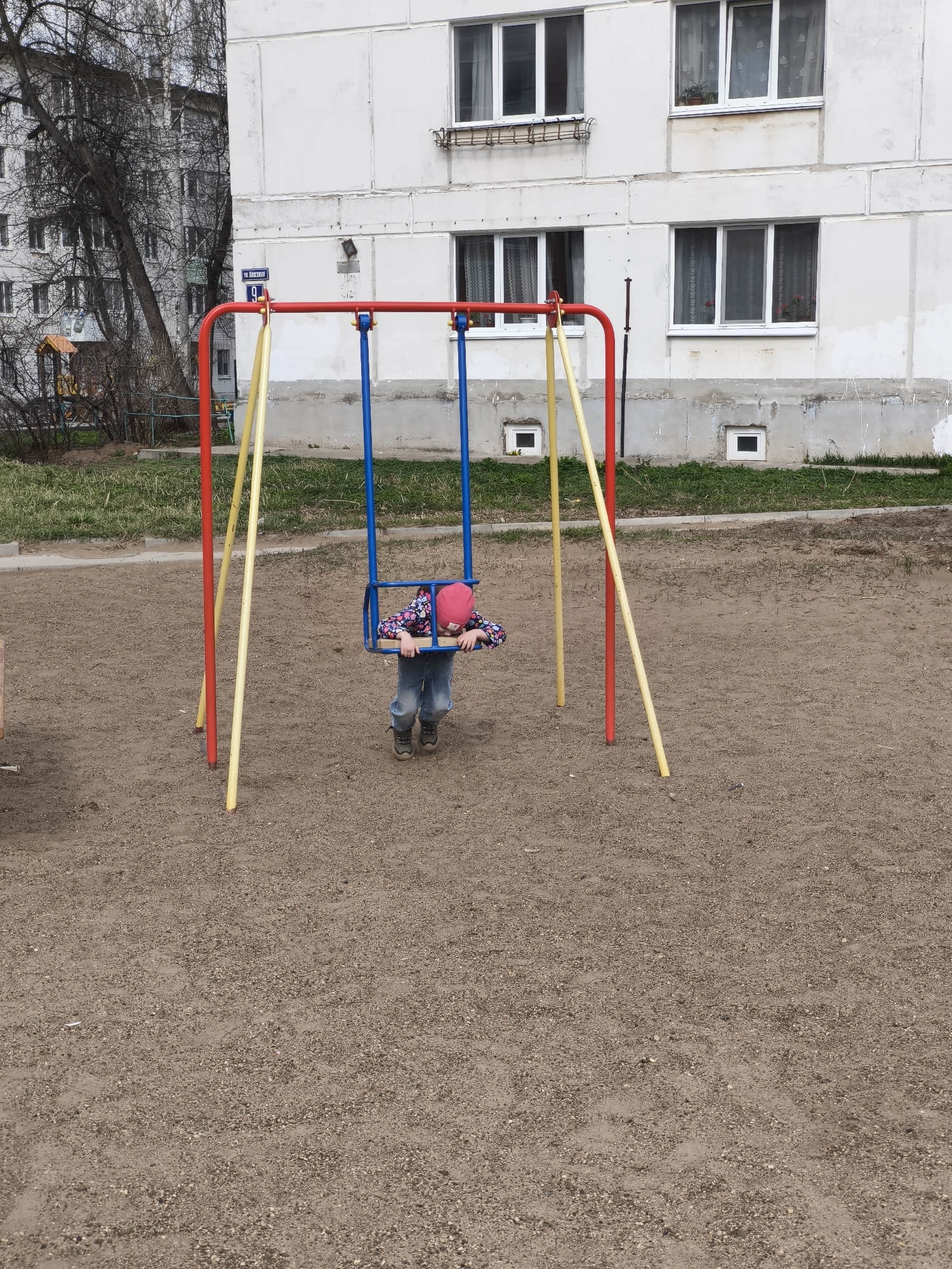 A low-angle, eye-level shot depicts a young child on a swing in a playground, with a multi-story building in the background. The child, clad in jeans, a colorful jacket, and a pink hat, appears to be resting her head on the swing's seat, holding onto the sides. The swing set is constructed with red horizontal bars, blue hanging chains, and yellow support poles.
The playground surface is covered in a brown gravel-like material. In the background, a light-colored building stands with multiple windows and a few plants in window boxes. A street sign reading "11 Druzgova" is visible on the left side of the frame near a tree. The environment suggests an urban setting, possibly a residential area with a communal playground.