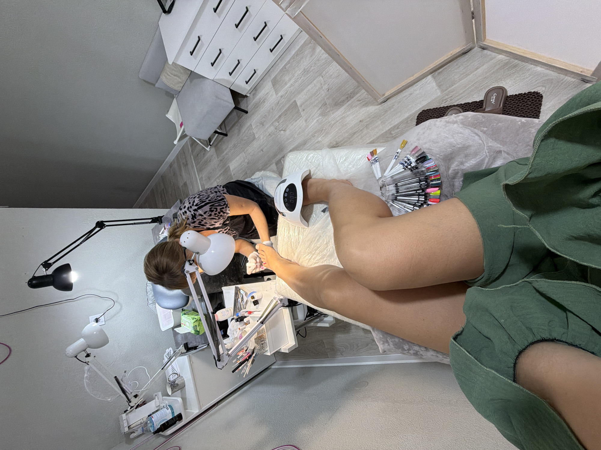 This is a forensic, matter-of-fact description of an image depicting a nail salon. The scene appears to be an indoor setting, likely a small salon or home-based business. A person, presumably a client, is seated with one leg extended forward, undergoing a pedicure. Their toenails are being worked on by a nail technician who is seated opposite them, wearing gloves and a leopard-print top. The technician is focused on the client's foot, holding a tool and likely applying nail polish or performing another treatment.
The environment includes a workstation with various nail care implements, bottles of polish, lamps, and other salon equipment. A UV nail lamp is positioned under the client's foot, indicating gel nail application. In the background, there is furniture, including a chest of drawers and a small armchair, suggesting a domestic or compact commercial space. The flooring is light-colored laminate. The lighting is provided by several adjustable lamps, casting focused illumination on the work area. There are also nail color swatches displayed on a rack. No specific geographical indicators are visible to pinpoint the location to Sochi, Russia, or any other specific city.