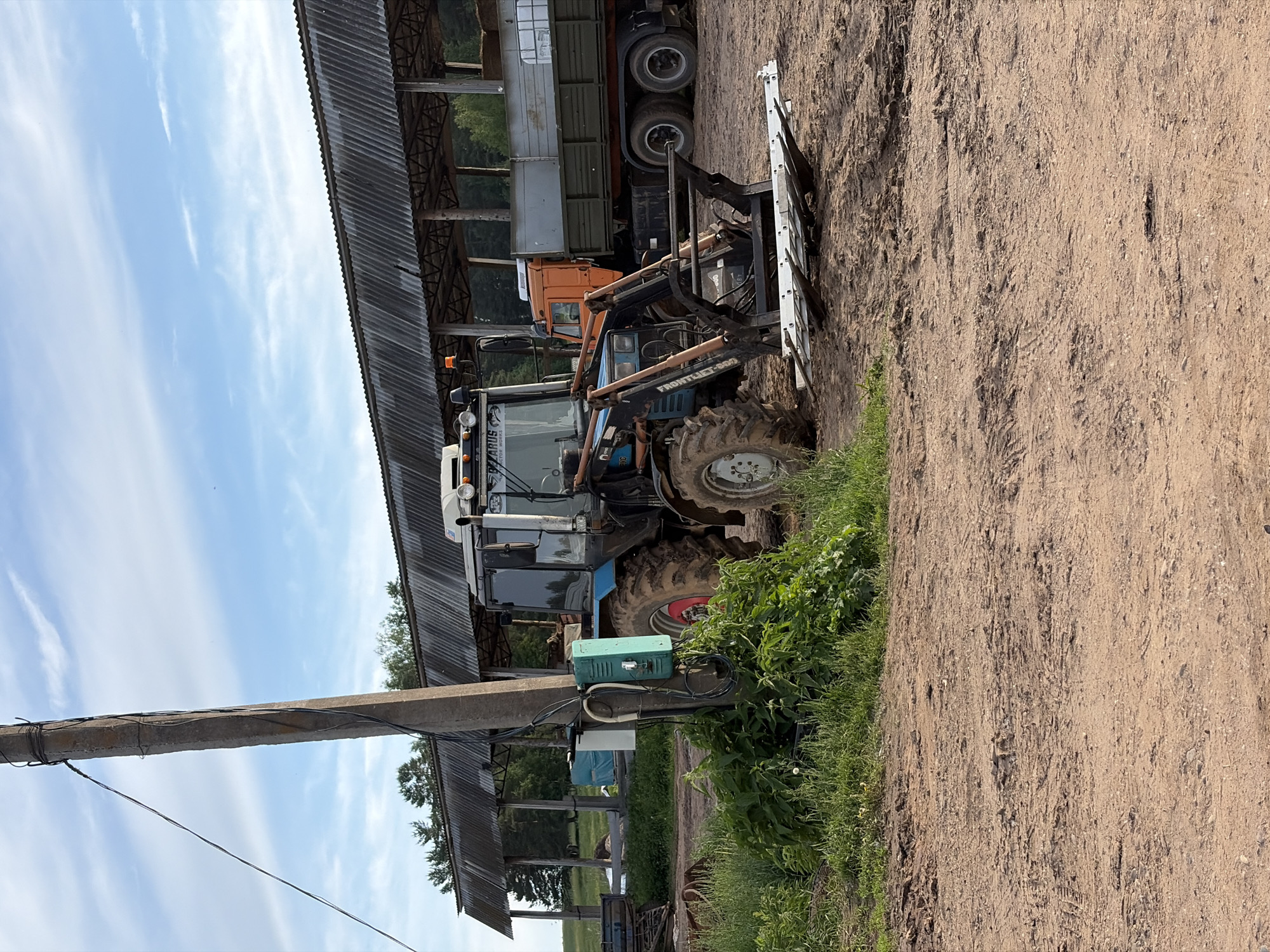 The image depicts a rural or agricultural setting in Чумна, Russia, under a bright blue sky with scattered wispy clouds. The scene features farm equipment parked alongside and partially within a long, open-sided shed with a corrugated metal roof.
In the foreground and to the right, a rough, earthy dirt path or ground is visible. To the left, a concrete utility pole stands, featuring several electrical wires and a light green junction box attached to it. Lush green vegetation, including grass and bushes, grows around the base of the pole and along the edge of the dirt area.
The primary subject is a blue and white tractor, likely a Belarus model, with "BELARUS" partially visible on the side. It is equipped with a front-end loader, which clearly displays the text "FRONTLIF P-812" on its arm. Another piece of text, possibly "BLG", is visible on the side of the engine compartment. The tractor's tires are muddy, suggesting recent use. Behind the tractor, an orange truck is partially visible, with a large, silver-sided trailer attached to it, both parked under the shelter of the shed.
In the background, beyond the shed, trees can be seen. The overall impression is a sunny day at a farm or industrial yard, with heavy machinery stored or temporarily parked. No specific activity is taking place; the scene is static.