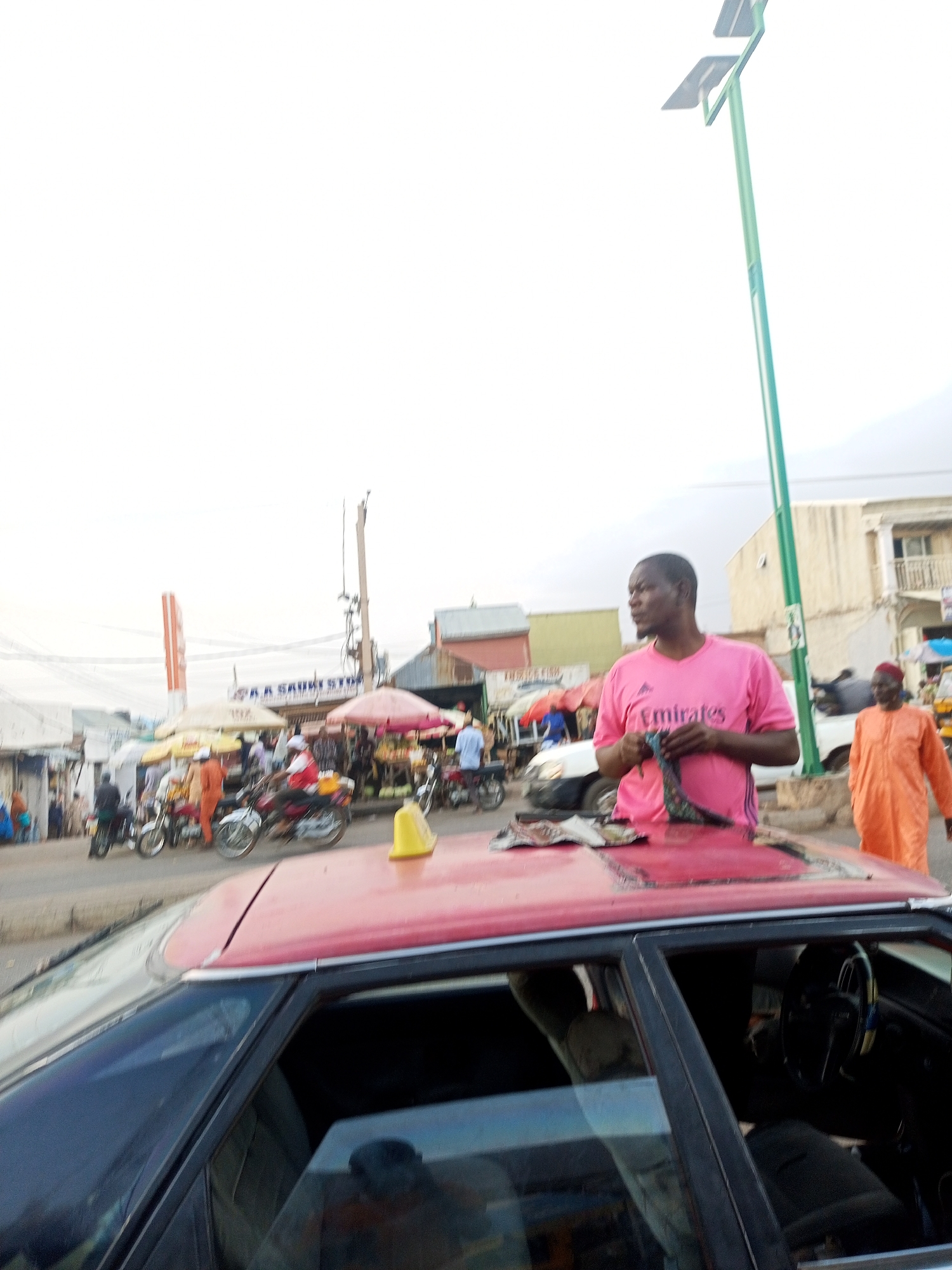 The image captures a street scene in Gombe, Nigeria, showcasing a vibrant and bustling market environment. In the foreground, a red car with a partially open sunroof dominates the view. A man in a pink shirt is leaning on the car, looking towards the right of the frame, and is holding what appears to be a piece of cloth. To his right, another man wearing an orange robe walks away from the camera.
The background is filled with a variety of subjects and objects indicative of a lively market. There are numerous people under parasols, many riding motorcycles, and storefronts with signs such as "A.A. SAUKI STORES." Overhead power lines are visible against a bright, cloudy sky. A green utility pole stands tall on the right.