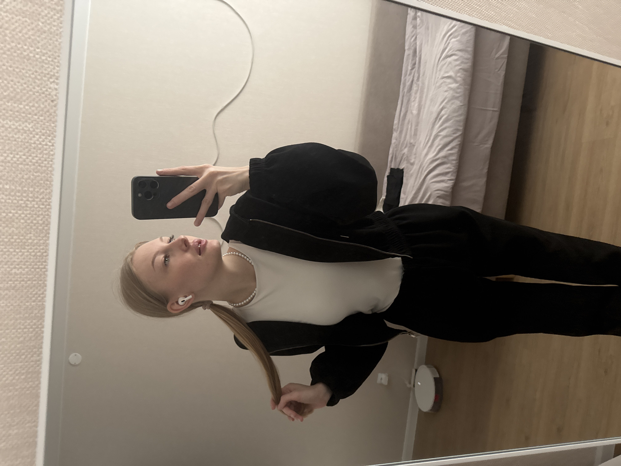 A young blonde woman is depicted in a mirror selfie taken within a room located in Perm, Russia. She is wearing a white fitted long-sleeve top, a black full-zip hoodie, and black pants. A pearl necklace is visible around her neck, and a white AirPod is present in her left ear. Her hair is styled into two pigtails, with her left hand gently grasping one. She holds a dark-colored smartphone with a prominent multi-lens camera system in her right hand, angled towards the mirror to capture the image. Her head is turned slightly to her right, and her gaze is directed away from the phone's camera.
The reflected environment reveals light beige walls and a light wood-toned floor. A textured, light-colored ceiling is visible at the top, featuring a small, circular white smoke detector. A white power cable extends horizontally across the wall in the background. A white robotic vacuum cleaner rests on the floor to the woman's left. A bed with a white duvet and light-colored fitted sheet is positioned in the background to her right, with a dark item draped over it near the foot. The mirror's frame is visible along the left and right edges of the composition.
