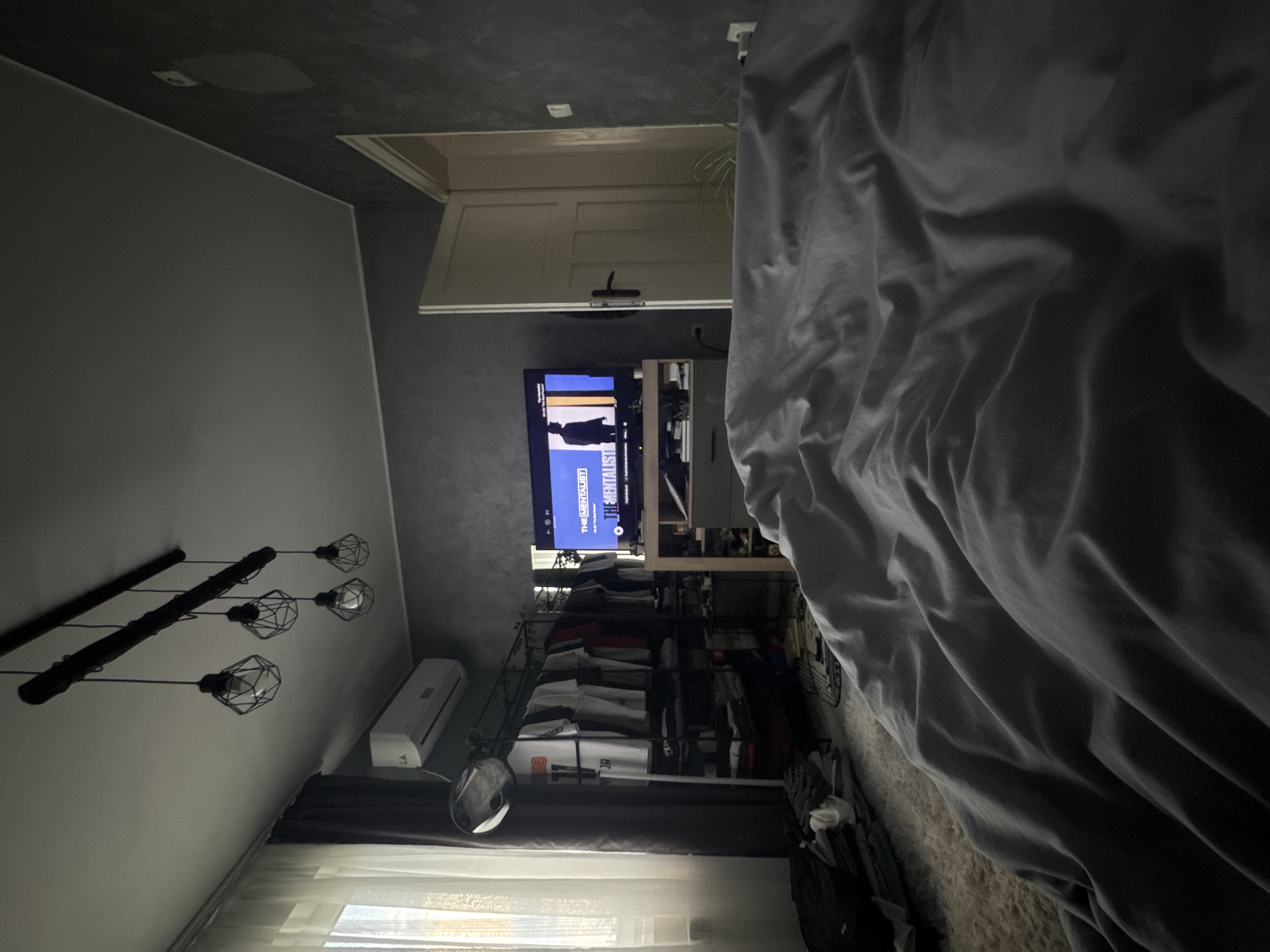 The image captures a dimly lit bedroom, viewed from the perspective of a bed partially visible on the right foreground with dark grey bedding. Directly across, a wall-mounted television displays the text "THE MENTALIST," indicating a show is playing. Below the TV is a dark, open shelving unit, and to its left, an open clothing rack filled with folded garments, with a small round mirror attached.
Above a window on the far left, a white wall-mounted air conditioning unit is visible. The window is covered by light-colored blinds or curtains, allowing some ambient light into the room. A distinctive light fixture featuring several bare bulbs enclosed in black geometric cages hangs from the dark grey ceiling. Above the television, a light-colored wooden door or cabinet is present. The walls are a neutral grey, providing a contrast to the darker ceiling. The scene appears to be a residential interior in Sofia, Bulgaria, characterized by modern furnishings and a relaxed atmosphere. No individuals are present in the image.