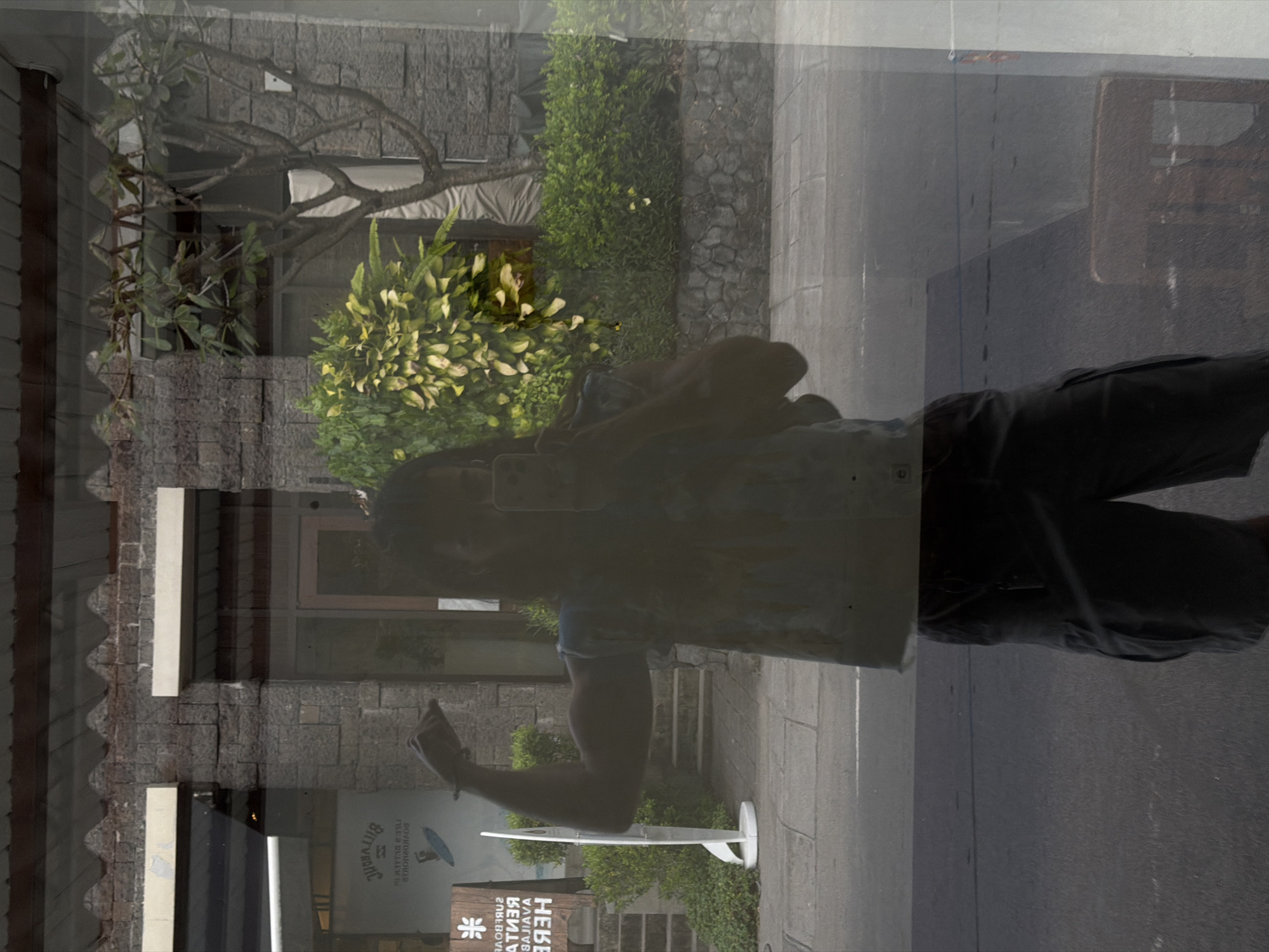 This media file presents a complex scene, primarily dominated by a reflection in a glass surface, likely a window, in North Kuta, Indonesia. The image is oriented vertically, rotated 90 degrees clockwise from its original capture.
The most prominent subject is the reflection of a person, appearing to take a selfie or record with a smartphone held in their right hand. The individual is wearing dark shorts and a light-colored, possibly patterned, short-sleeved top. Their form is partially transparent and blended with the background due to the reflective nature of the glass.
In the reflected foreground, a white surfboard stands upright on a display stand. Behind it, a sign with reversed text is visible, which reads "SURFBOARD RENTAL AVAILABLE HERE." Below this, a logo resembling a snowflake or asterisk is present, alongside partially visible text that appears to be "BINTANG" with a red star logo, indicative of a common Indonesian brand.
The background, viewed through the glass and intertwined with the reflection, reveals stone-walled buildings and lush tropical vegetation. Various green bushes and trees, including one with distinctive gnarled, bare branches, are visible. The architecture suggests a local style. The ground appears to be paved, possibly a sidewalk or courtyard. The bright illumination indicates it is daytime with clear weather. Faint reflections of other structures, possibly a chair or table, can be seen in the glass to the right.