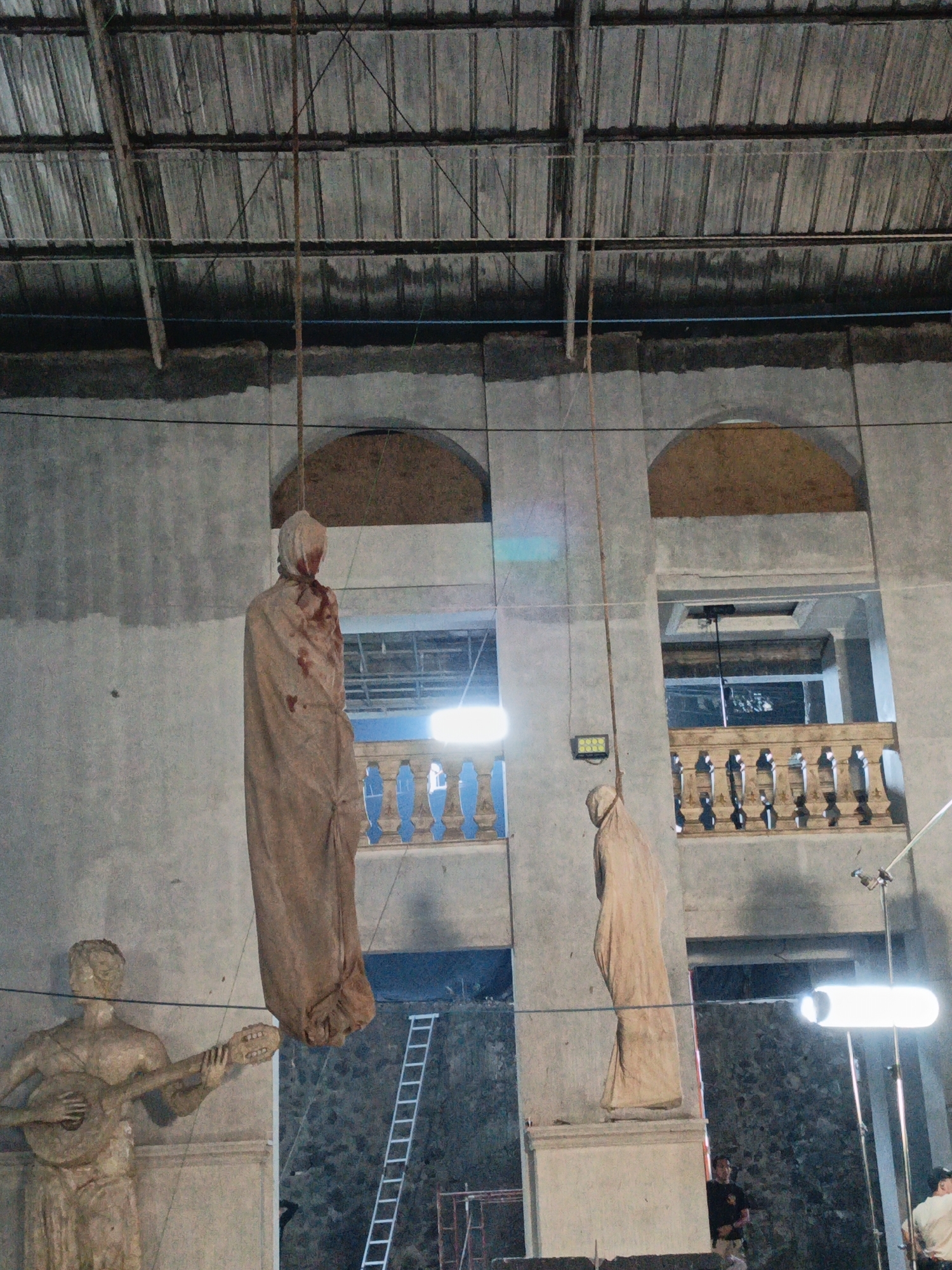 An eerie scene unfolds within a dimly lit, cavernous interior, possibly an abandoned building or a set constructed for a production in Cibogo, Indonesia. The raw concrete walls and corrugated metal ceiling suggest an unfinished or industrial space. Two shrouded figures, resembling mannequins or props, hang suspended by ropes from the ceiling. The figure on the left is draped in a tan cloth stained with what appears to be blood, particularly around the head. The second figure, also draped in tan fabric, hangs slightly to the right and lower than the first.
In the foreground, a stone statue of a man playing a lute stands on a pedestal. To the right of this statue, a ladder leans against a stone wall. A balcony with ornate railings is visible in the background, with two arched openings above it. Lighting equipment, including a bright, horizontal light fixture, is positioned to the right of the scene, suggesting an active filming or photography session. Two individuals are partially visible in the background; one man in a dark shirt and khaki pants stands near the balcony, and another man in a light-colored shirt is partially visible on the far right. The overall atmosphere is somber and unsettling, with the hanging figures and dim lighting creating a sense of suspense or despair.