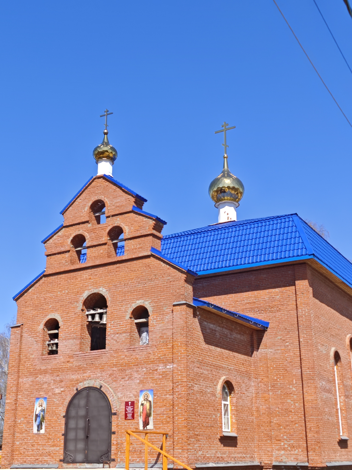 The image captures a brick church in the city of Фоки, Russia. The church is constructed from red bricks and has a blue tiled roof. It features two golden domes, each topped with a cross. The church has a bell tower with arched openings where bells are visible. The front of the church has a dark metal door, flanked by two icon paintings and a small sign. The sky is a clear, bright blue. A yellow railing is visible in the foreground.