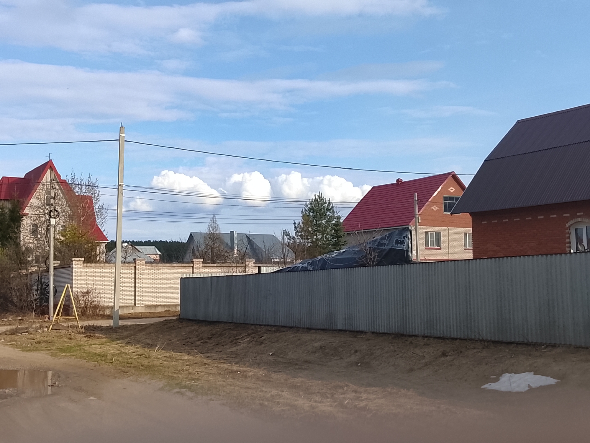 The image depicts a street scene in Chaikovsky, Russia. A gray corrugated metal fence runs horizontally across the middle ground, separating the street from a row of houses. The houses are varied in style and color, with red and dark gray roofs and brick and siding exteriors. Brick fences separate the houses. Power lines stretch across the sky, passing in front of cumulus clouds. The foreground consists of an unpaved dirt road with puddles. A yellow A-frame sign is visible near a power pole on the left side of the image. Patches of grass and bare earth are visible along the roadside. A small patch of snow lies on the ground near the right edge of the frame.
