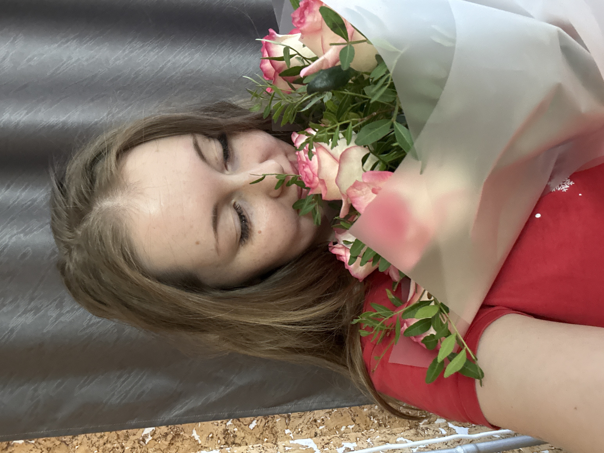 A woman with light brown hair and closed eyes is holding a bouquet of pink and white roses wrapped in translucent white paper. She is wearing a red shirt. The background is a gray textured surface, and a lower section with a cork-like texture is visible. The location is not identifiable as Unknown, Unknown.
