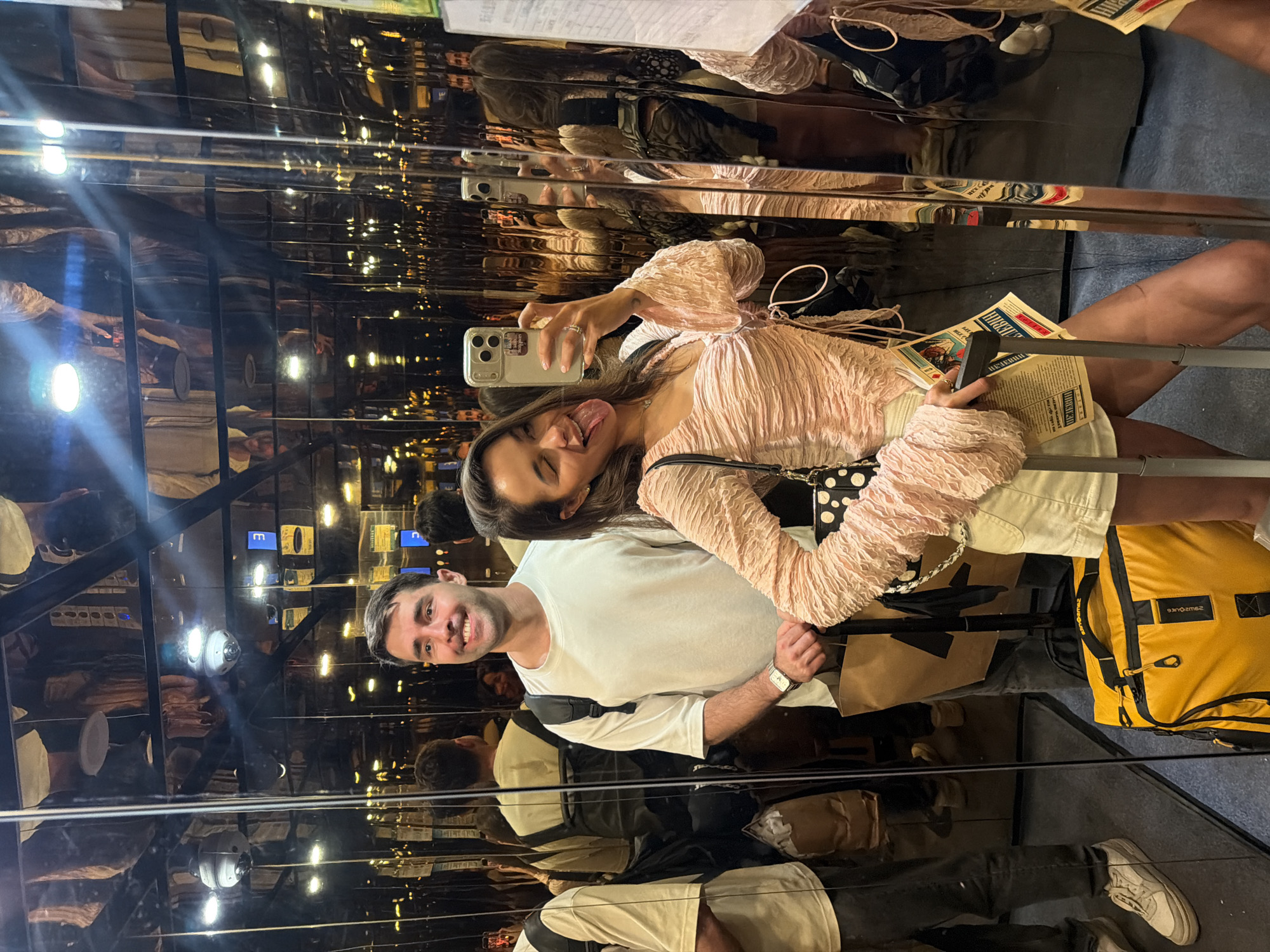 A man and a woman are taking a selfie in an elevator lined with reflective surfaces. The woman is holding a smartphone and sticking her tongue out. The man is smiling. They are surrounded by reflections of themselves and the elevator interior. There is luggage on the floor, including a yellow backpack. There are newspaper objects in the woman's arms and on the man's shoulder. The location is not discernible as Zhangjiajie, China from the image.
