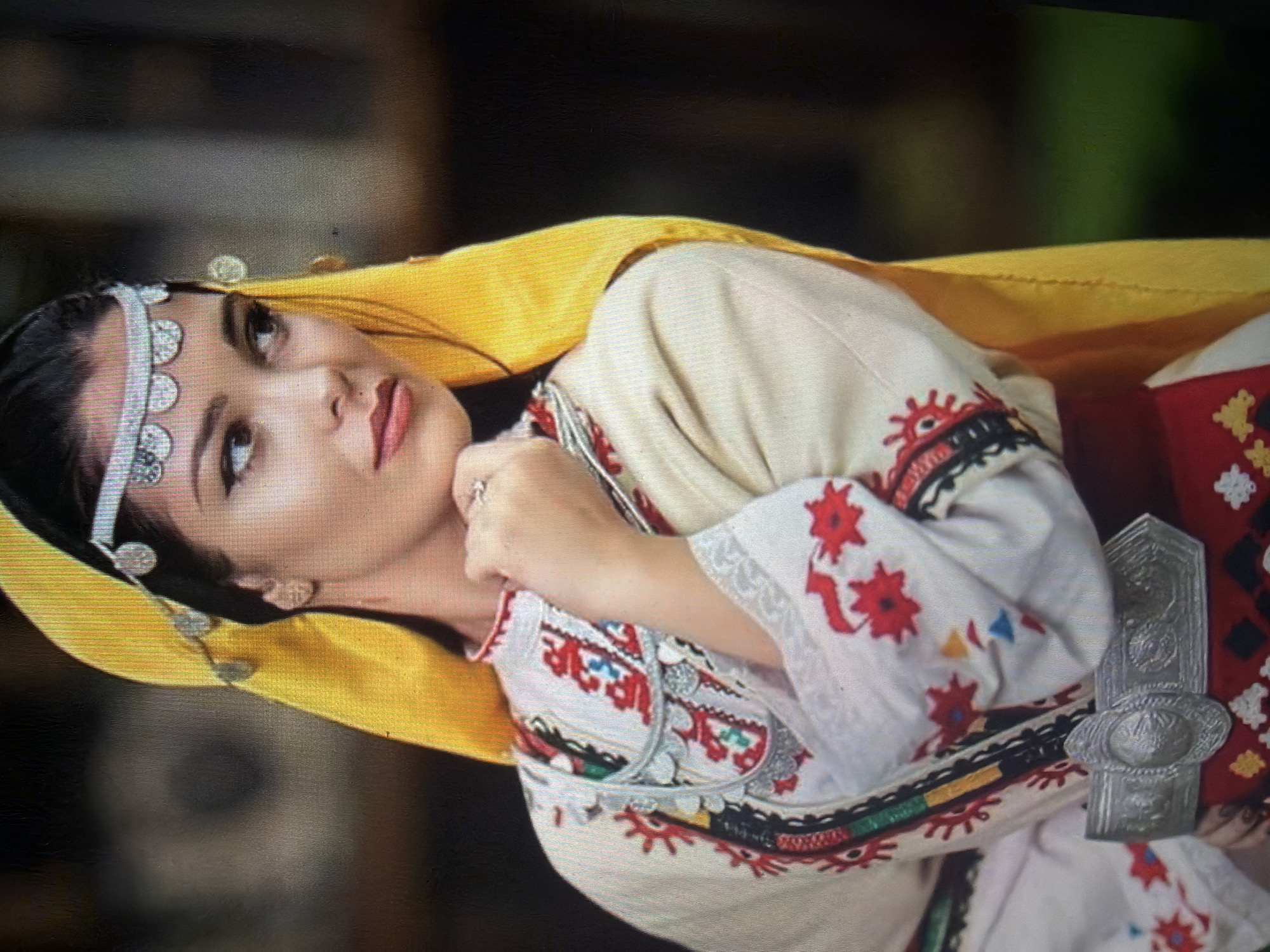 The image features a female subject depicted from the upper torso, oriented at a slight angle towards the viewer's left, with her gaze directed to the right of the frame. She wears a bright yellow headscarf partially covering dark hair, over which a silver-toned headpiece with a band of semi-circular disc ornaments is positioned across her forehead. Smaller silver, leaf-shaped ornaments are also present on the headscarf near her right temple. Her face shows dark eyes with winged eyeliner, light pink lipstick, and a visible nose piercing on her right nostril. She is adorned in a traditional garment, a long-sleeved cream or off-white blouse, featuring extensive folk embroidery. This embroidery includes red floral or star-shaped motifs on the sleeves and chest, complemented by geometric patterns in red, black, blue, and yellow on vertical decorative bands. A silver-toned decorative chain, incorporating smaller disc-like elements, is visible around her neck. Her right hand, wearing a ring on the ring finger, is raised with fingertips resting near her chin. A wide, ornate silver metal belt, characterized by a central oval buckle and embossed patterns, secures the attire at the waist, overlying a red lower garment portion that displays white and yellow geometric embroidery. The background is indistinct, out of focus, and predominantly dark, consistent with an outdoor setting in the city of Shumen, Bulgaria.