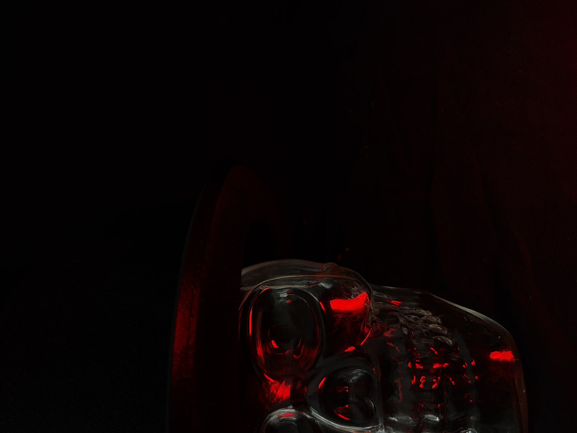 A close-up, low-angle shot features the upper portion of a clear, glass skull illuminated by a bright red light. The skull is positioned on the left side of the frame, with its facial features clearly defined. A black hat is visible resting on top of the skull. The background is predominantly dark, with some faint red lighting present on the right side of the image, creating a contrasting and eerie atmosphere. The scene is devoid of any people, actions, or interactions. The environment is dark and appears to be an indoor setting with minimal distractions.