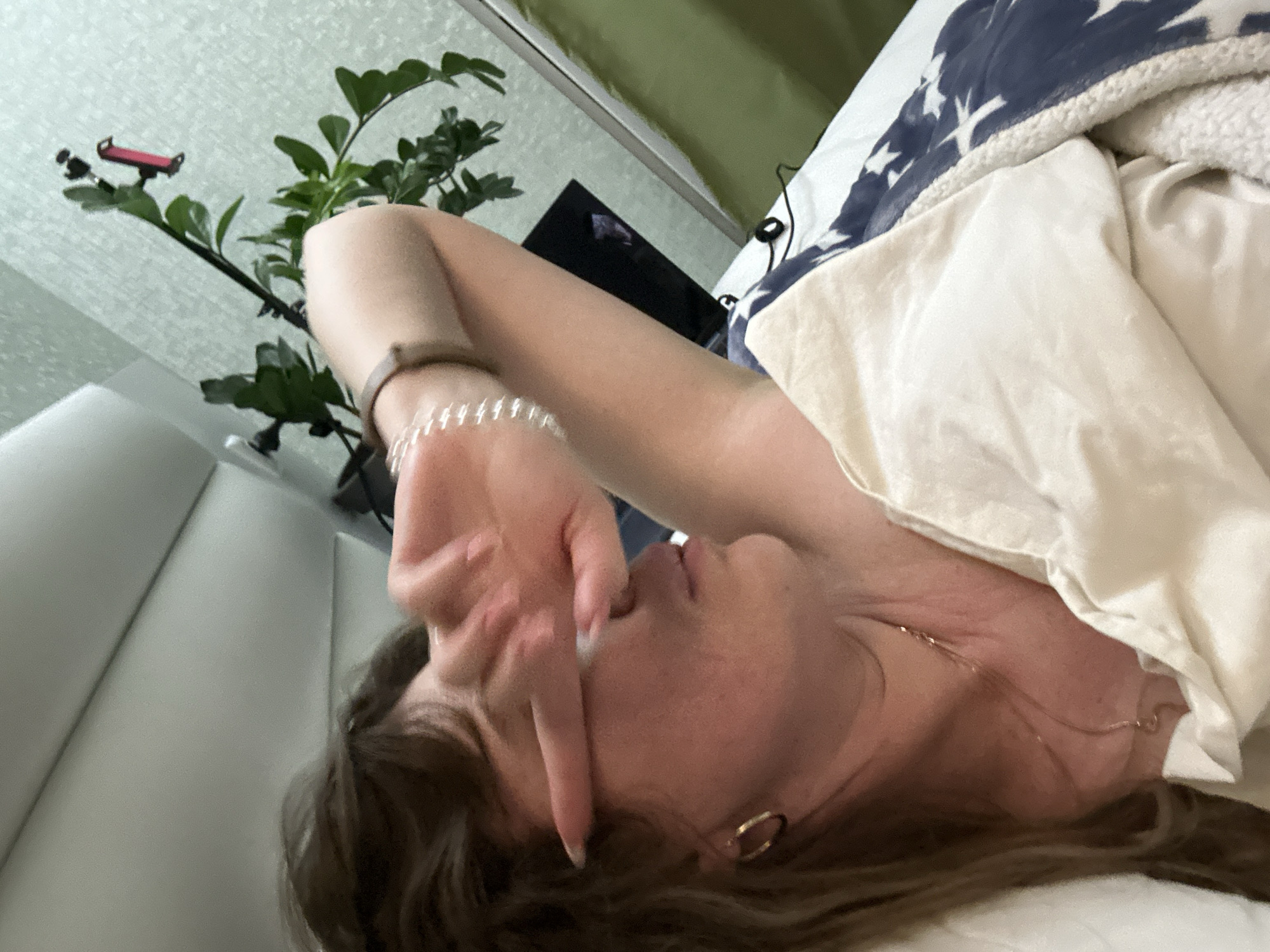A woman is lying in bed, with her right hand covering her eyes. She is wearing bracelets on her right wrist and a gold necklace. The bed has white and blue patterned bedding. A green plant is visible to the left of the frame, along with a black television. The location is a bedroom and is not indicated in Batumi, Georgia.