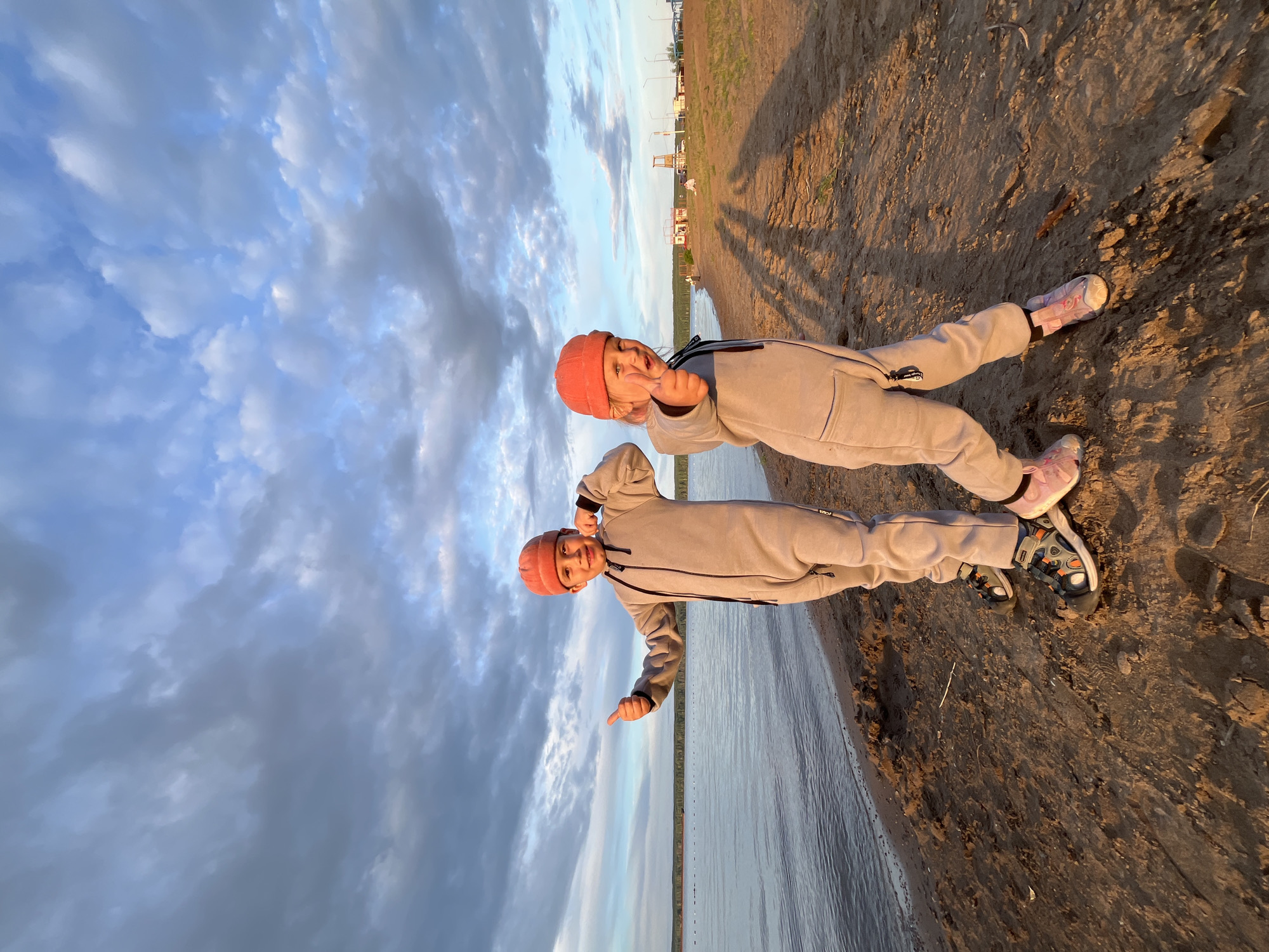 The image shows two young children lying on a sandy embankment near a body of water, possibly a lake or river. The children are wearing matching light gray jumpsuits and orange beanies. The child in the foreground, on the right, is looking up at the camera with a playful expression, holding up a finger to their mouth. The other child, to the left, is also looking towards the camera.
The sky above is filled with dramatic clouds, suggesting either early morning or late evening. The sun is low in the sky, casting long shadows on the sand, which indicates it's either sunrise or sunset. The water is calm and reflects the sky. In the background, on the horizon, there are some buildings and structures, possibly indicating a populated area or a resort.
The overall mood of the image is cheerful and relaxed, with the children appearing to be enjoying their time outdoors. The location is identified as Chaikovsky, Russia. No visible text is present in the image.
