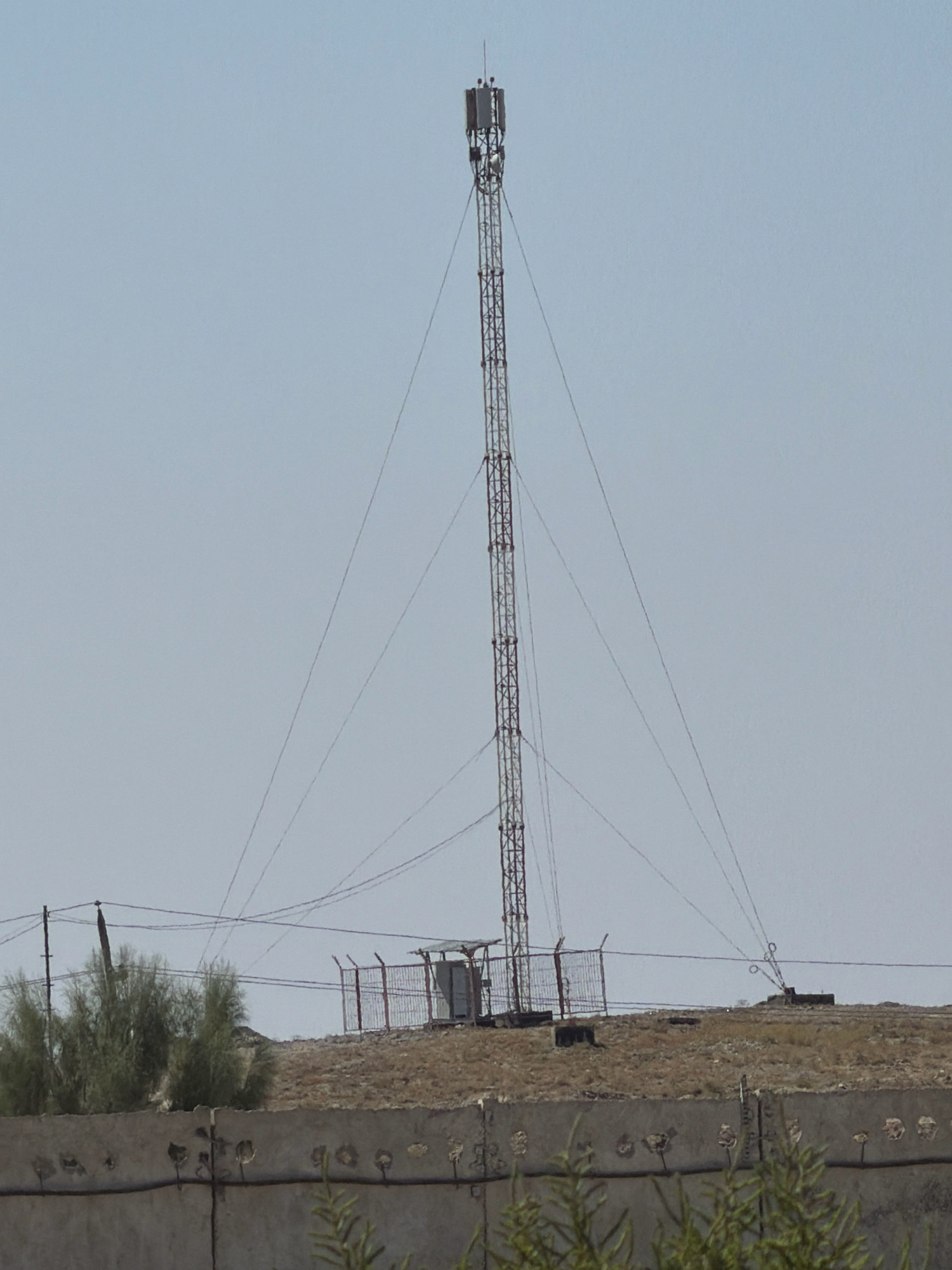 This is a medium-distance, horizontal, eye-level shot of a cell tower and its surrounding area on a clear, overcast day. The sky is a pale blue. The cell tower is a tall, lattice-structure metal tower with antennas at the top. It is supported by guy wires. To the left of the tower is a fenced-off area containing a small, shed-like structure. In the foreground, there is a concrete wall with holes and some greenery visible. The ground around the tower is dry and brown, with scattered vegetation. The overall impression is one of an industrial or telecommunications site in a rural or semi-rural environment. The location is Besapan, Uzbekistan.