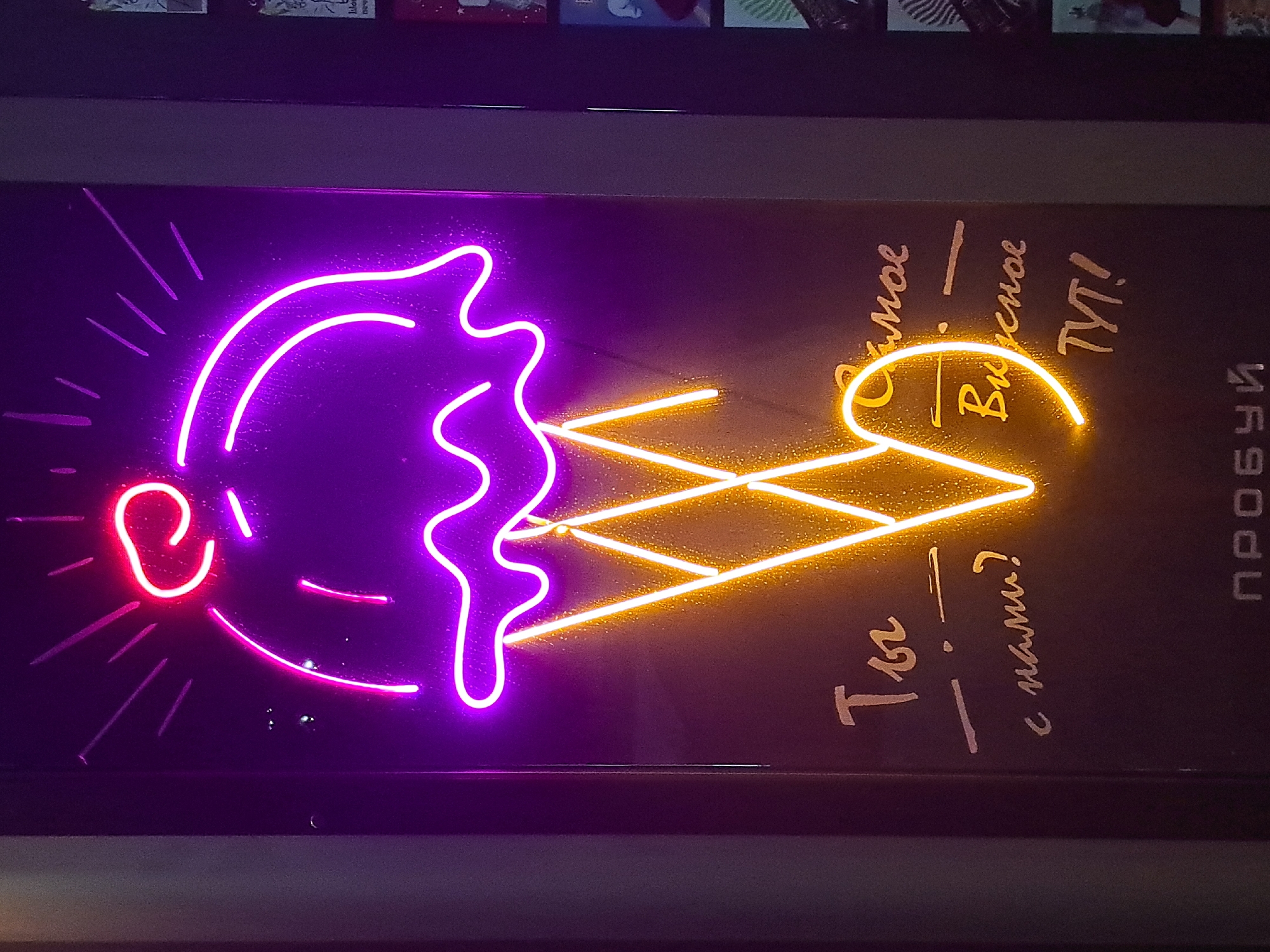 An ice cream cone is depicted in neon lights, primarily in purple and orange hues. The ice cream itself is represented by a curved purple shape with small red accents at the top, suggesting a cherry or topping, and thin purple lines emanating outwards, possibly indicating sparks or flavor. The cone is outlined in bright orange neon. Below the cone, in a handwritten-style font, are the Russian words "Ты — с нами?" which translates to "You—with us?". To the right of this, also in a similar font and the same orange neon, is the phrase "Самое вкусное тут!" meaning "The tastiest is here!". At the very bottom of the image, in a simpler block font, is the word "ПРОБУЙ", meaning "TRY". The neon sign is mounted on a dark, matte surface, possibly a wall or a display board. The lighting creates a vibrant and inviting atmosphere. The image was taken indoors.