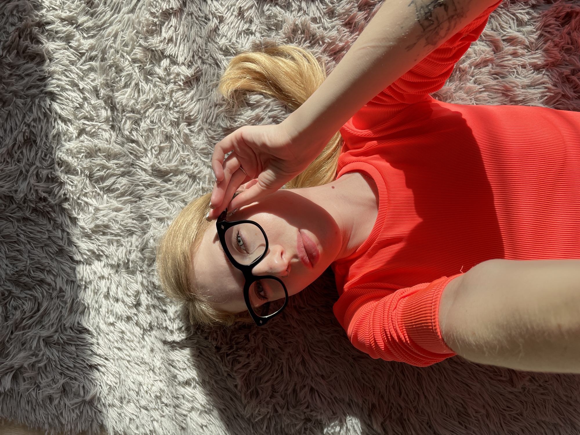 A woman with blonde hair tied in a ponytail is lying on a plush, light grey rug, possibly indoors. She wears a bright orange, ribbed long-sleeved shirt and black-rimmed glasses. One hand is positioned near her face, seemingly adjusting her glasses. Her left arm, showing a tattoo, extends upwards, while her right arm extends downwards. The background consists solely of the rug, which is partially illuminated by sunlight, creating a contrast of light and shadow. The city of Izhevsk, Russia, is not discernible from the provided image.