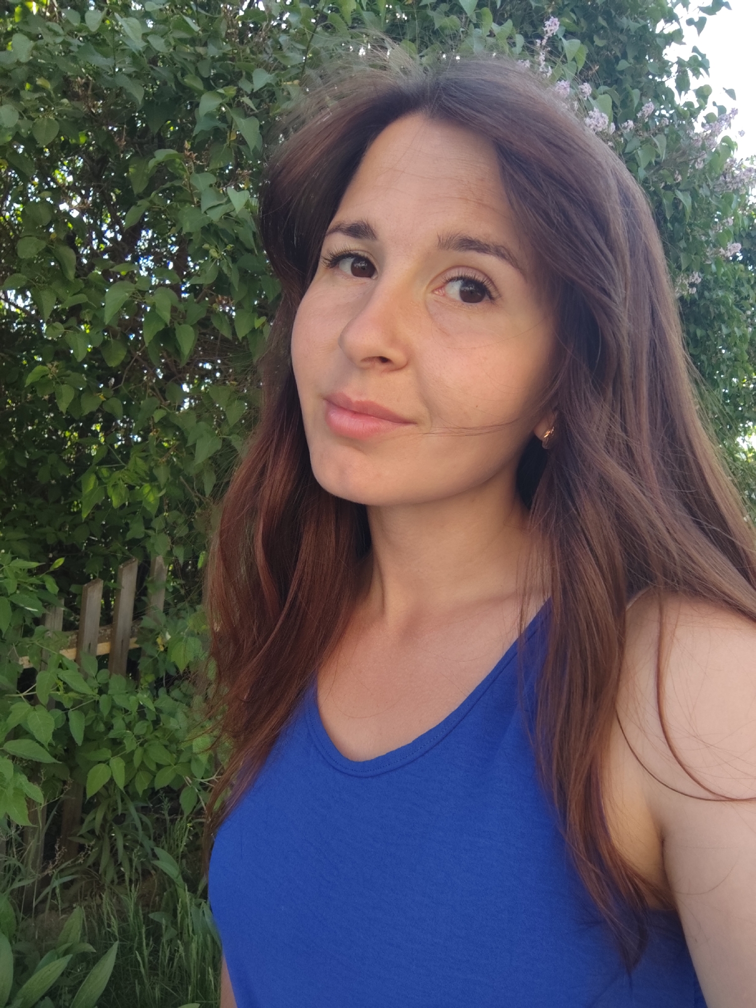 A woman with long, auburn hair and light skin is centered in the image, facing the camera with a slight smile. She is wearing a blue tank top. Behind her, lush greenery fills the background, with trees and some foliage visible. To the left, a partially visible wooden fence adds depth to the scene. The overall setting appears to be outdoors, possibly in a garden or park. The location in the city of Фоки, Russia cannot be determined from this single image.