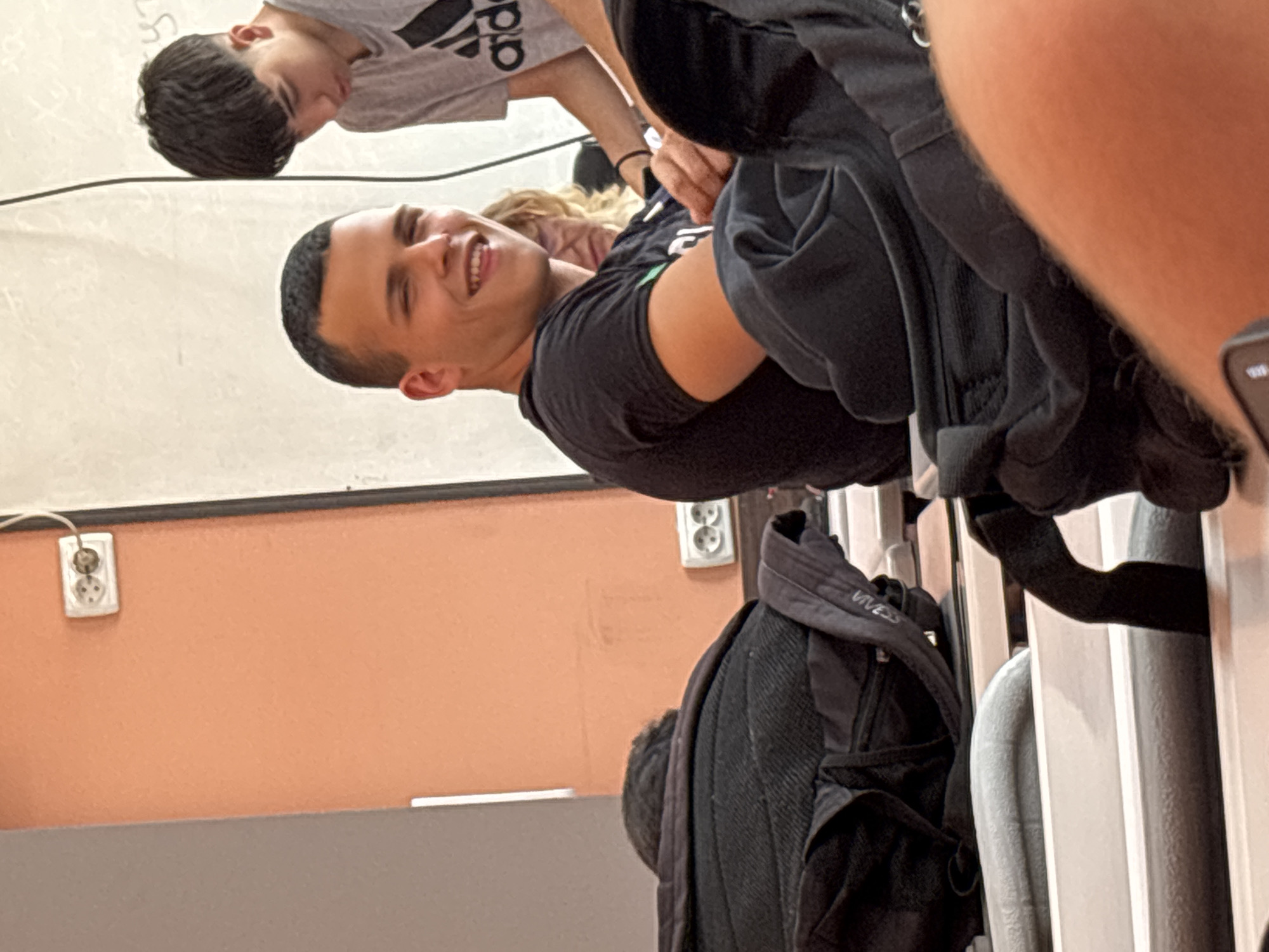 The image, originally rotated 90 degrees counter-clockwise, shows an indoor scene featuring several young individuals. The main subject, a young man with short dark hair, is smiling broadly at the camera. He wears a black short-sleeved shirt with a green design visible on his left sleeve. Behind him, a person with blonde or light brown curly hair is partially visible. Further in the background, another young man with dark, wavy hair, wearing a gray t-shirt with a partially visible "adidas" logo, is looking downwards and away from the camera.
The setting appears to be a casual indoor space, possibly a classroom or common area. The background includes a light-colored wall, an orange-painted wall section, and a dark brown or black cable running horizontally. A white electrical outlet is visible on the orange wall. In the lower right and bottom of the frame, several black and gray backpacks or bags are hung on what seem to be chairs or a railing. One black bag clearly shows the text "VIVES". Faint, partially obscured text (possibly "LS") is written on the light-colored wall in the background. The smiling expression of the central figure suggests a positive and relaxed mood. The time of day is not discernible, as the lighting appears to be entirely artificial.