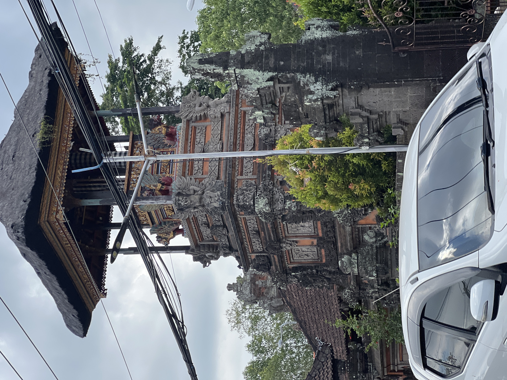 A white car is parked in front of a traditional Balinese temple entrance in Abiansemal, Indonesia. The temple features intricate stone carvings and a thatched roof. The sky is overcast, and there are many overhead power lines crossing the image. Lush green trees and vegetation surround the temple. The car's windshield reflects the cloudy sky and surrounding structures.