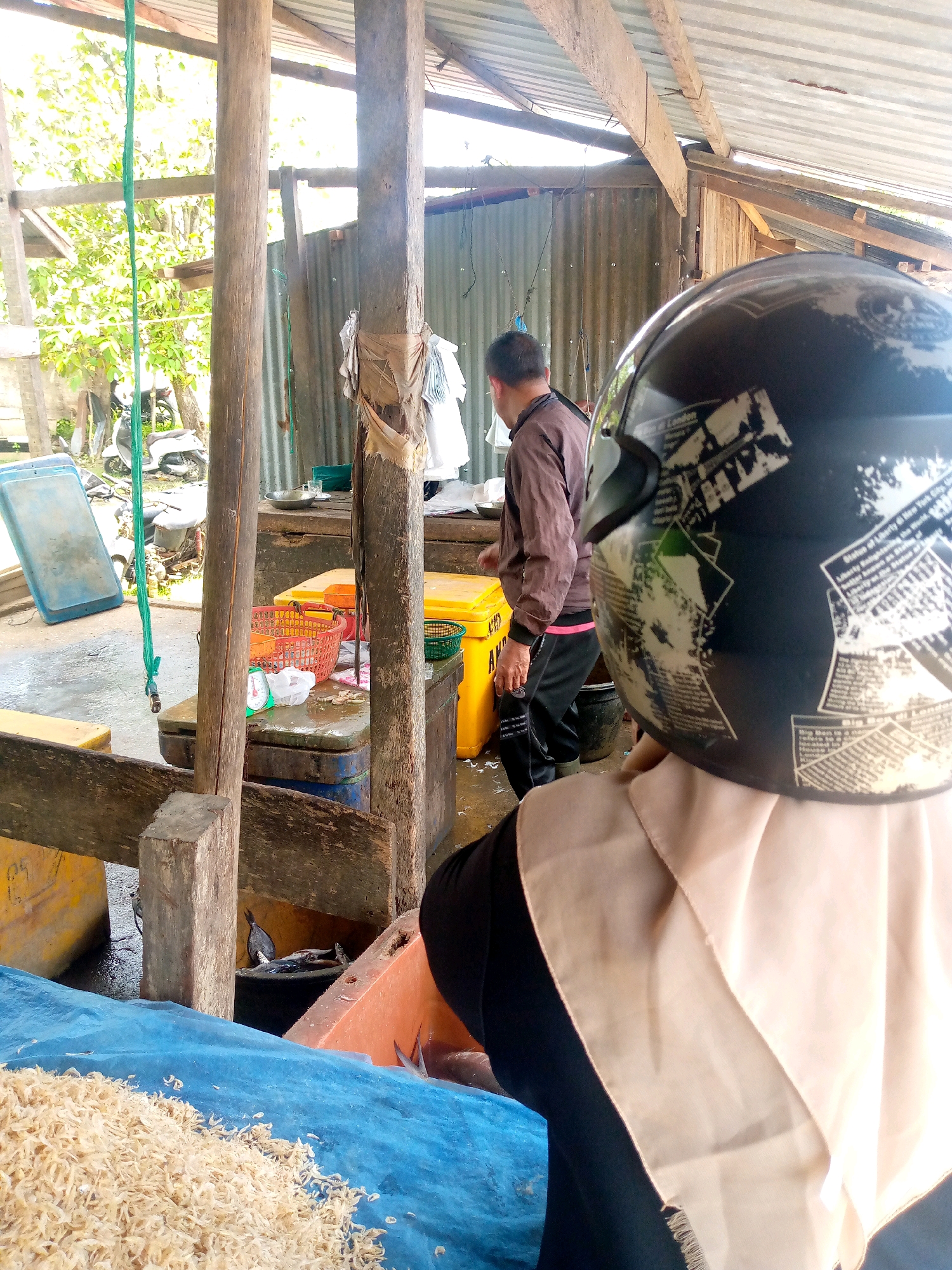This image depicts a market scene in Mancang, Indonesia. In the foreground, the back of a person wearing a black helmet with newspaper-like decals and a light brown hijab is visible. In the middle ground, a man in a brown jacket and dark pants is seen standing in front of a yellow cooler and a red basket containing fish. Several other fish are also visible in tubs and on a table. To the left, a blue plastic container and a green rope hang from wooden posts. A pile of dried shrimp is spread on a blue tarp in the lower left corner. The background reveals a rustic market structure with a corrugated metal roof, wooden pillars, and more market stalls. The environment suggests an outdoor or semi-outdoor market, likely focused on the sale of fresh and dried seafood.