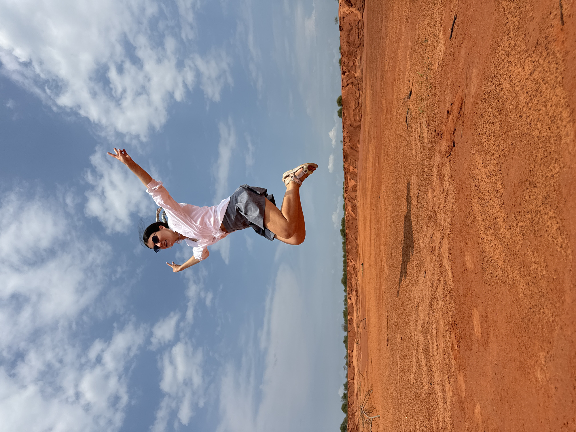 A young woman with dark hair, wearing sunglasses, a white collared shirt, and a gray skirt, is captured mid-air as she jumps against a blue sky with white clouds. She is in a desert-like landscape with bright orange-red sand, indicative of the red sand dunes in Mui Ne, Vietnam. The ground is uneven with scattered dry branches and vegetation. The woman's arms are raised, with her right arm extended forward and her left arm bent, both hands open. Her legs are bent at the knees, with her right foot forward and her left foot trailing. The scene depicts a moment of joy and freedom in a natural, arid environment.