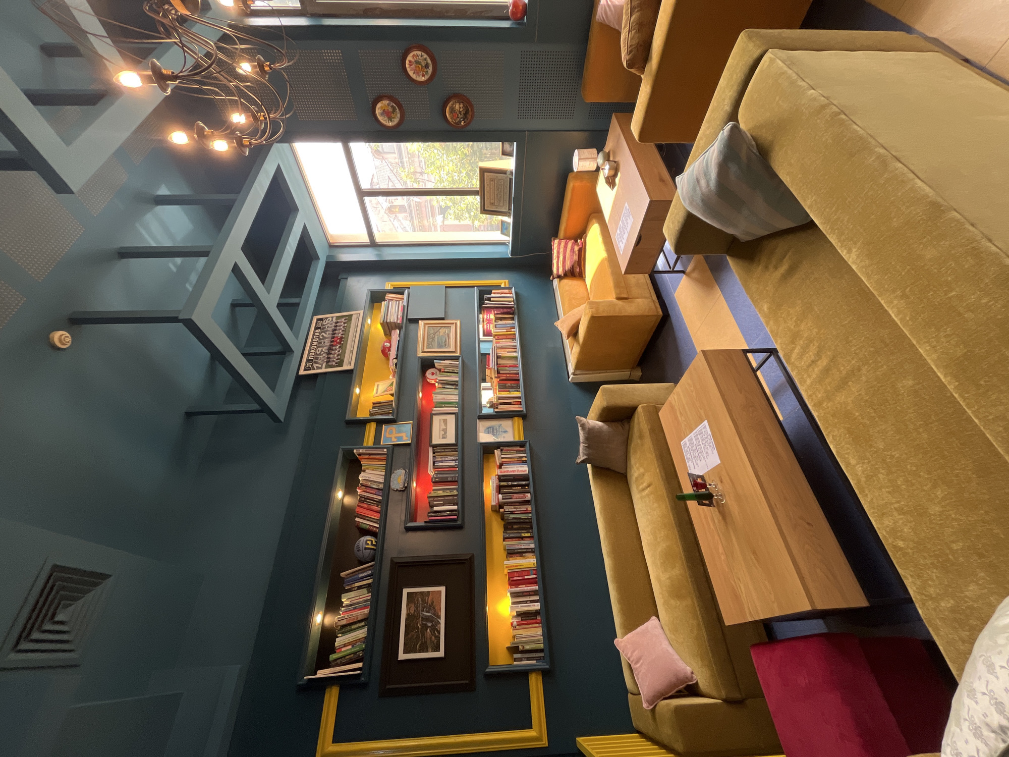 The image showcases the inviting and stylish interior of a lounge or cafe, characterized by its deep teal walls and mustard-yellow furnishings. Natural daylight streams in through a large window, suggesting it is daytime, and is supplemented by artificial lighting from a prominent multi-bulb chandelier and integrated lights within the wall-mounted bookshelves.
The room is furnished with several plush, mustard-yellow sofas and armchairs, arranged around low wooden coffee tables. Scattered cushions, including a light blue striped one and a soft pink one, add to the comfort. A deep red upholstered stool is visible under one of the tables.
The teal walls are a central design element, featuring geometric structural accents that extend from the walls and ceiling. They are tastefully decorated with small, round, floral-patterned plates and an eclectic collection of irregularly shaped bookshelves. These shelves are filled with numerous books and some are illuminated from within, creating a warm glow. Various framed artworks, including a large black-framed picture and smaller pieces, are interspersed among the books. A bold yellow frame highlights a section of the bookshelf wall.
No people are present, indicating a quiet moment in the establishment. The atmosphere appears cozy, relaxed, and conducive to reading, conversation, or quiet contemplation. While there is visible text on a paper on one of the coffee tables and a framed sign near the window, the details are illegible. The design, with its blend of warm colors and decorative details, creates an artistic and comfortable environment.