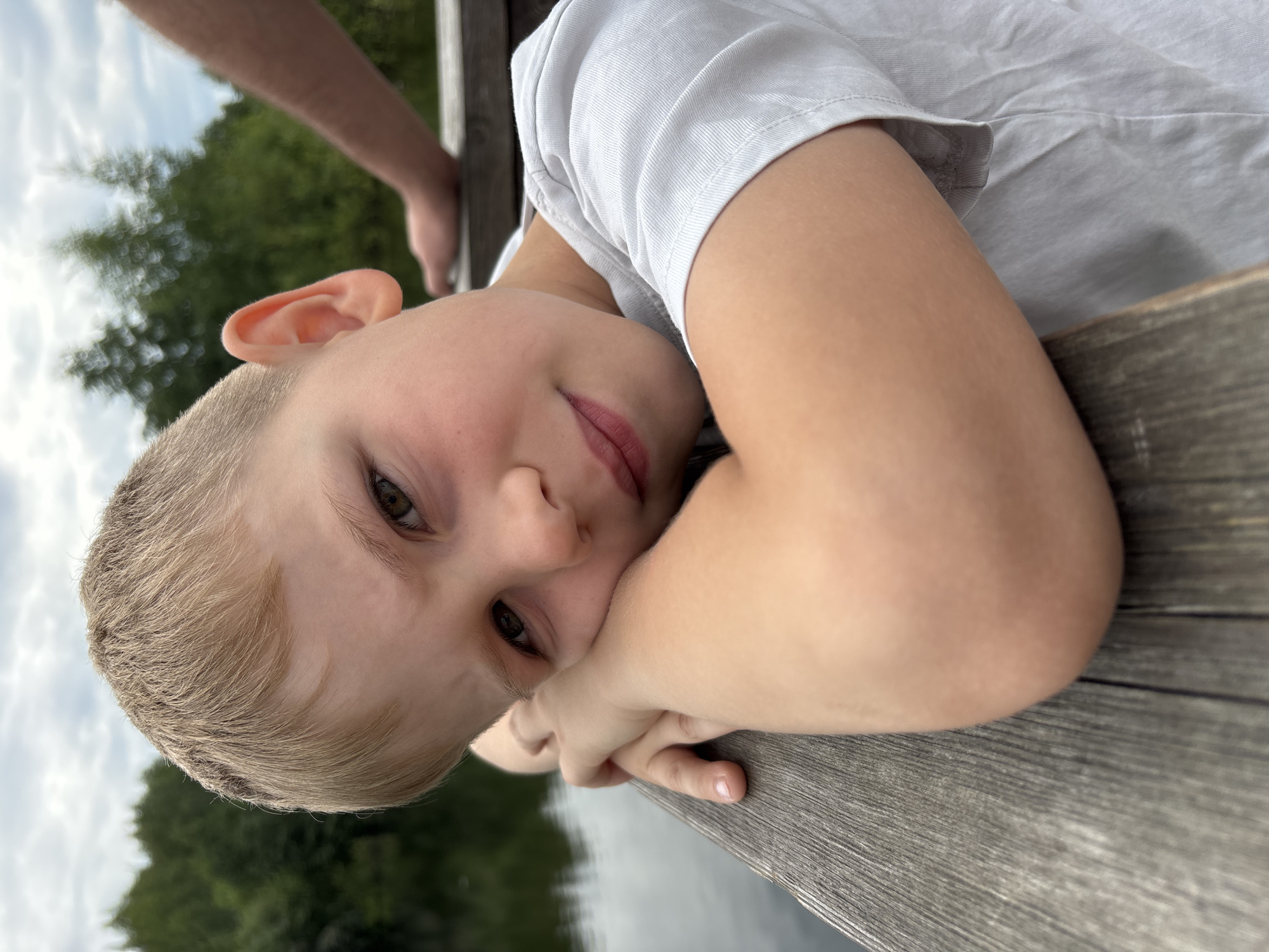 A close-up, eye-level medium shot captures a young Caucasian boy with short blond hair leaning on a weathered wooden railing. His left arm is propped up, supporting his head as he looks directly at the camera. His eyes are wide and brown, and his lips are slightly parted in a neutral expression, with subtle pink lipstick. He is wearing a light gray t-shirt. The background is blurred, showing a body of water to the left and a line of green trees against a cloudy sky. To the right, a portion of another person's arm and hand is visible, also resting on the railing. The overall setting appears to be outdoors, likely near a lake or river. There is no discernible indication of the location being within the city of Yaroslavl, Russia.