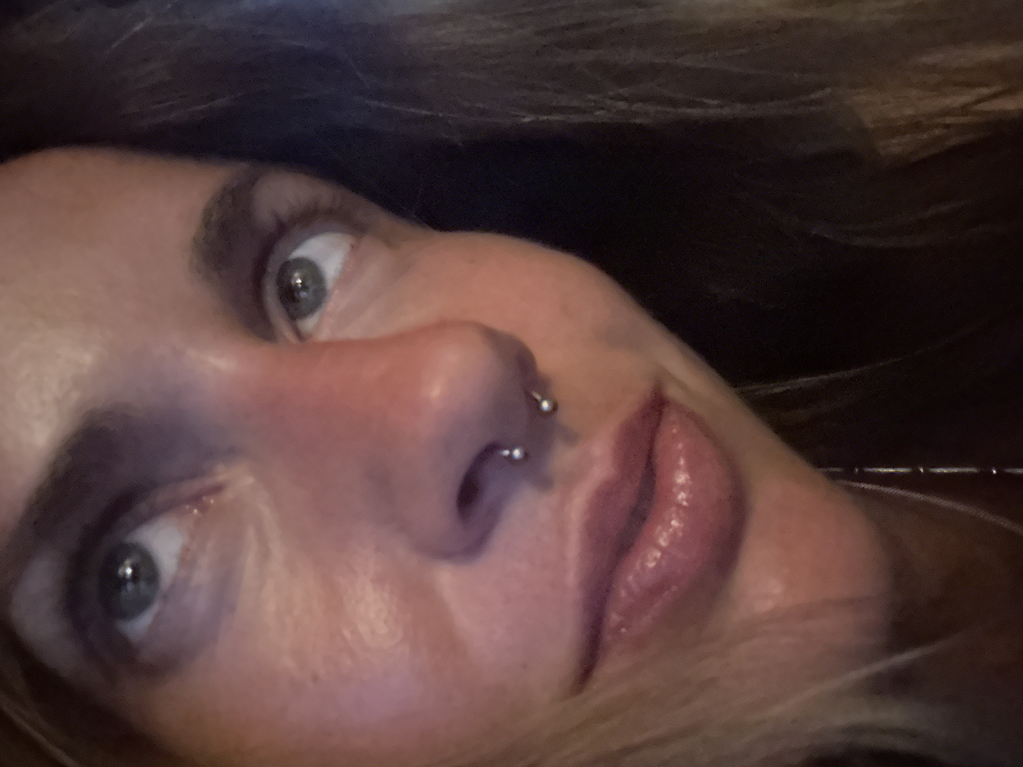 A close-up, slightly blurry, eye-level, candid photograph captures the upper half of a fair-skinned woman's face, angled towards the right. Her gaze is directed upwards and to the left. The lighting is warm, highlighting her skin with a rosy hue, particularly on her cheeks and nose. She has blue-green eyes, visible eyebrows, and full lips with a subtle sheen of gloss. A septum piercing, consisting of a circular barbell with two silver balls, is visible on her nose. Her blonde hair is blurred and frames the right side of her face. The background is dark and indistinct, suggesting an indoor setting. There is no discernible urban environment or specific location.