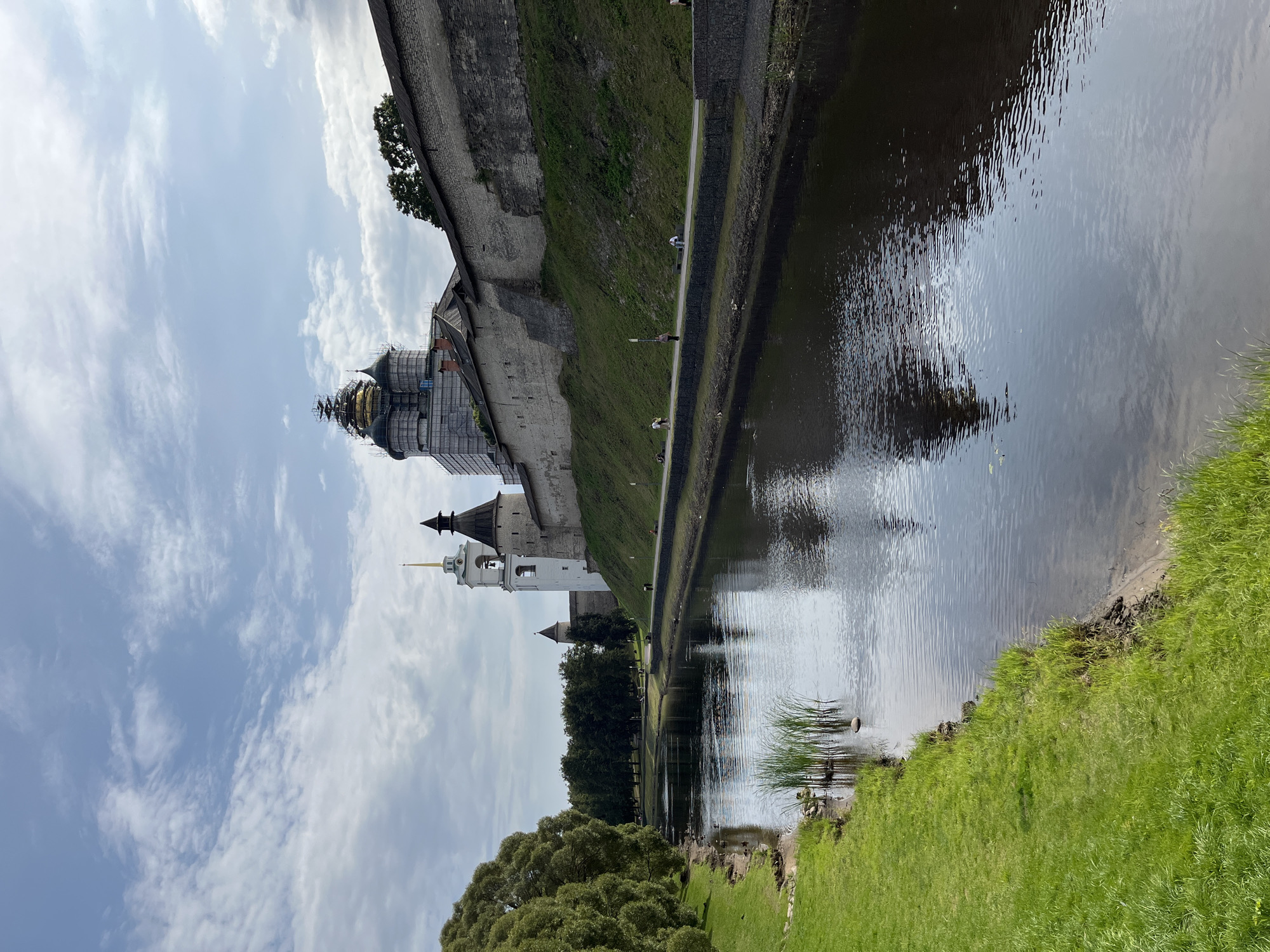 The image depicts the Pskov Krom (Kremlin), a historic fortress located in Pskov, Russia. The Krom is situated on a high promontory at the confluence of the Pskov and Velikaya rivers. In the foreground, the Velikaya River flows calmly, reflecting the cloudy sky and the surrounding greenery. The left bank of the river is covered with lush green grass and trees.
On the right side of the river, the imposing stone walls of the Krom rise up, with grassy slopes leading to them. Several towers of the Krom are visible, including the distinctive Trinity Cathedral with its golden dome and a white bell tower. Adjacent to it, a darker, conical-topped tower stands. Further along the wall, scaffolding is visible around a structure undergoing renovation, possibly another dome or tower.
A paved walkway runs along the base of the Krom walls, parallel to the river. Several individuals are seen walking along this path, some sitting on benches. Ducks are visible in the water near the grassy bank on the left. The sky above is partly cloudy, with a mix of white and grey clouds. The overall scene is one of historical grandeur and tranquil natural beauty.