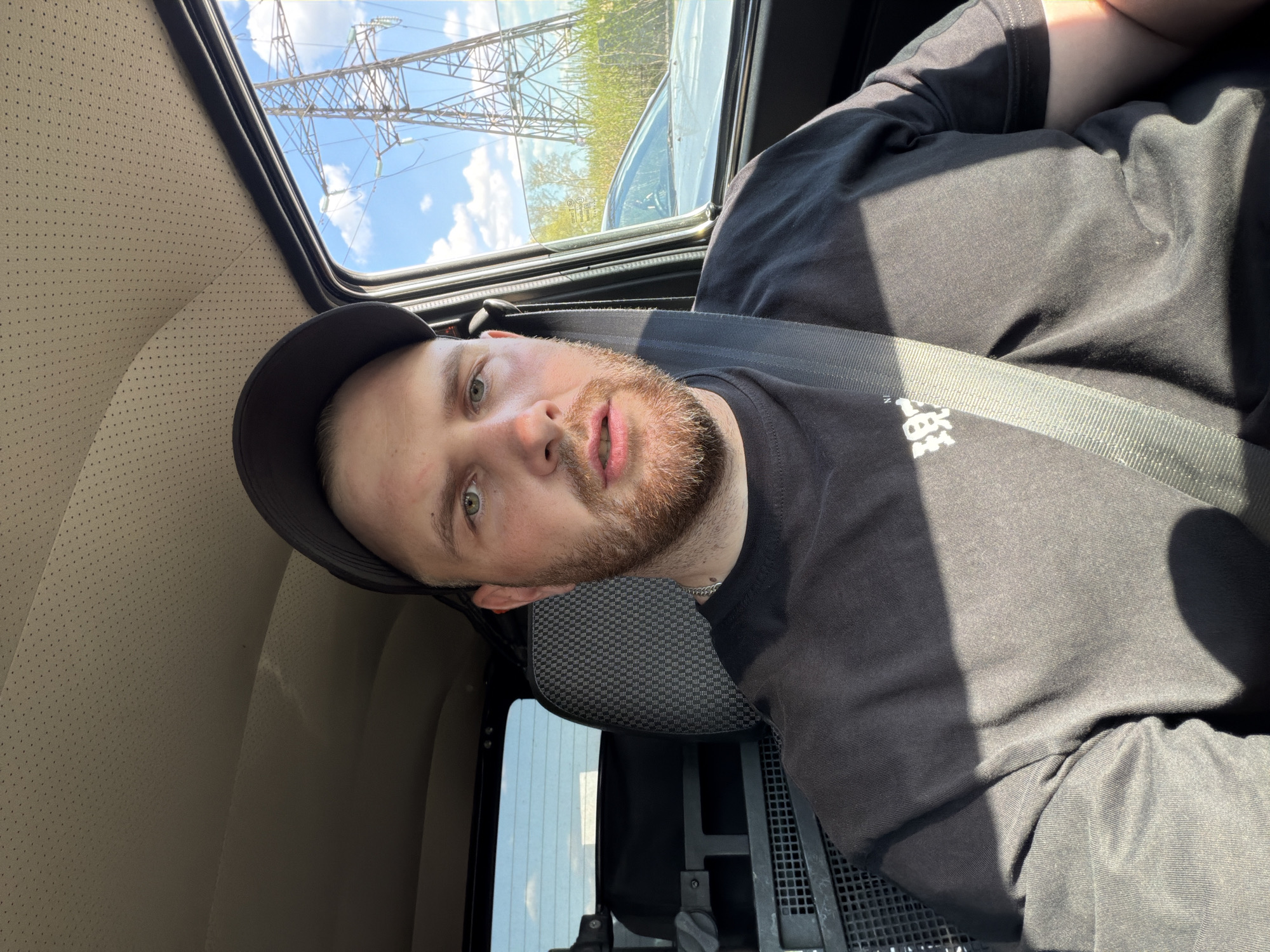 A bearded man wearing a black cap and a black t-shirt is sitting inside a vehicle wearing a seatbelt. Outside the window, there are electricity pylons. The vehicle appears to be in motion. The location may be Chaikovsky, Russia.
