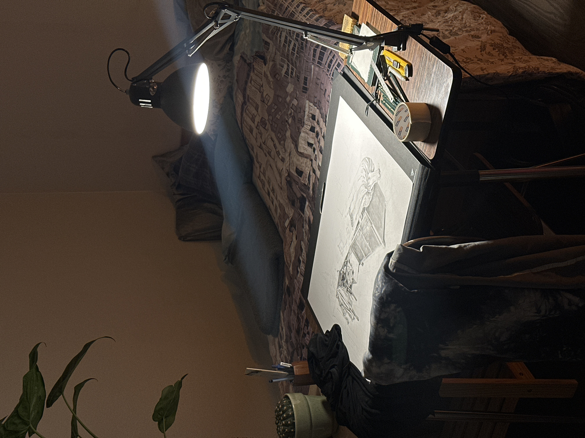 The image depicts a drawing setup indoors. A black articulated lamp is positioned above a drawing board, illuminating a work-in-progress drawing of a figure on white paper. The drawing board rests on what appears to be a bed, partially covered by patterned bedding. Various drawing tools and supplies, including pencils, a ruler, and masking tape, are placed on the board alongside the drawing. A cactus in a pale green pot is visible in the lower foreground, along with plant leaves. The room is dimly lit, with the primary light source being the lamp. The location is not identifiable as Montreuil, France, based on the visible contents of the image.