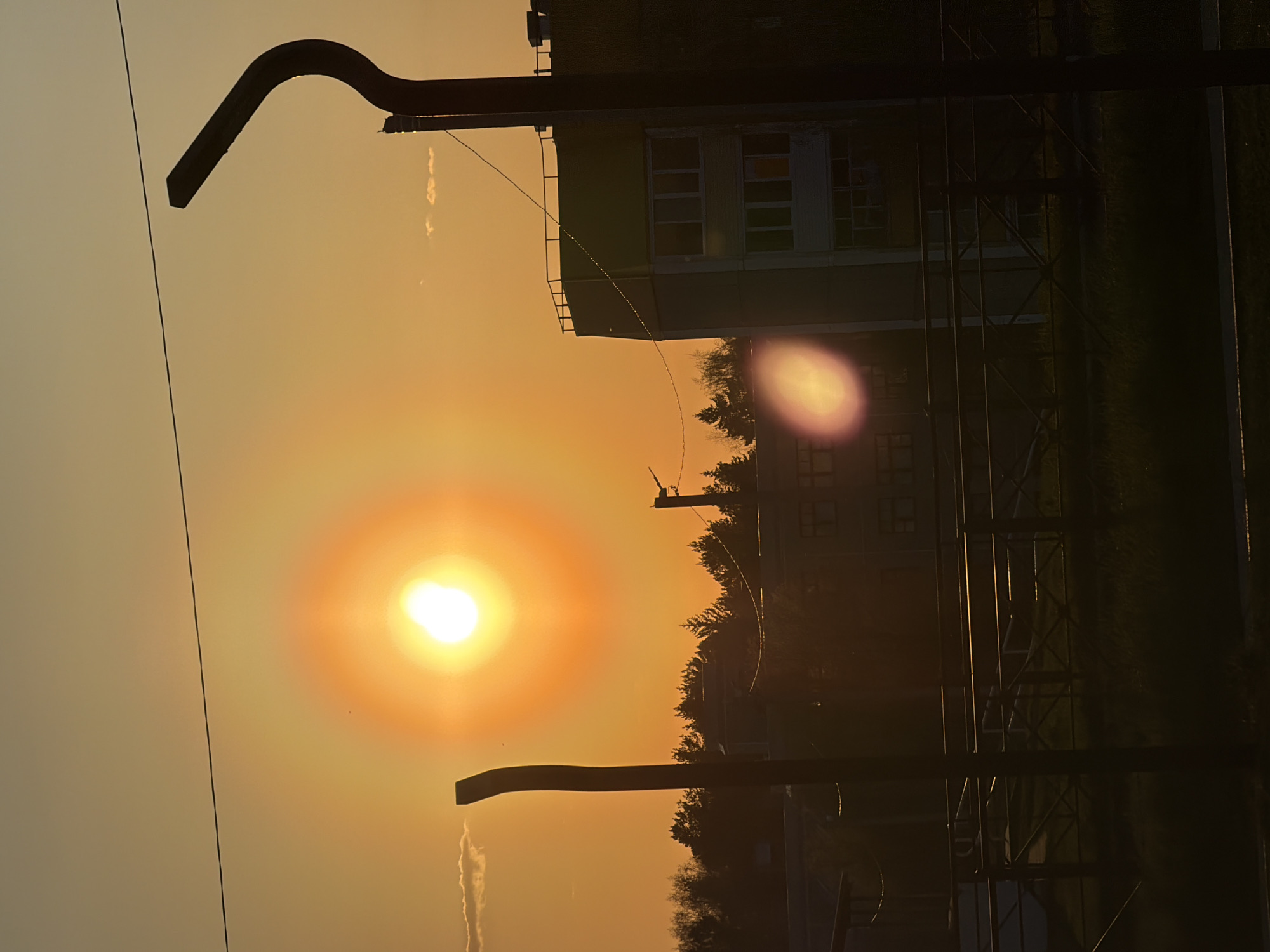The image depicts a sunset scene in Марковский, Russia. In the foreground, two curved black metal structures resembling street lamps are visible. A power line cuts through the upper left of the frame. The sun, encircled by a bright orange halo, is prominent in the center of the image. Behind the street lamps and power lines, a dark green building with white-framed windows is partially visible. A row of trees runs along the horizon. The general lighting suggests dusk or dawn, casting long shadows and emphasizing silhouettes. The overall impression is a hazy, golden-hued cityscape under a setting or rising sun.