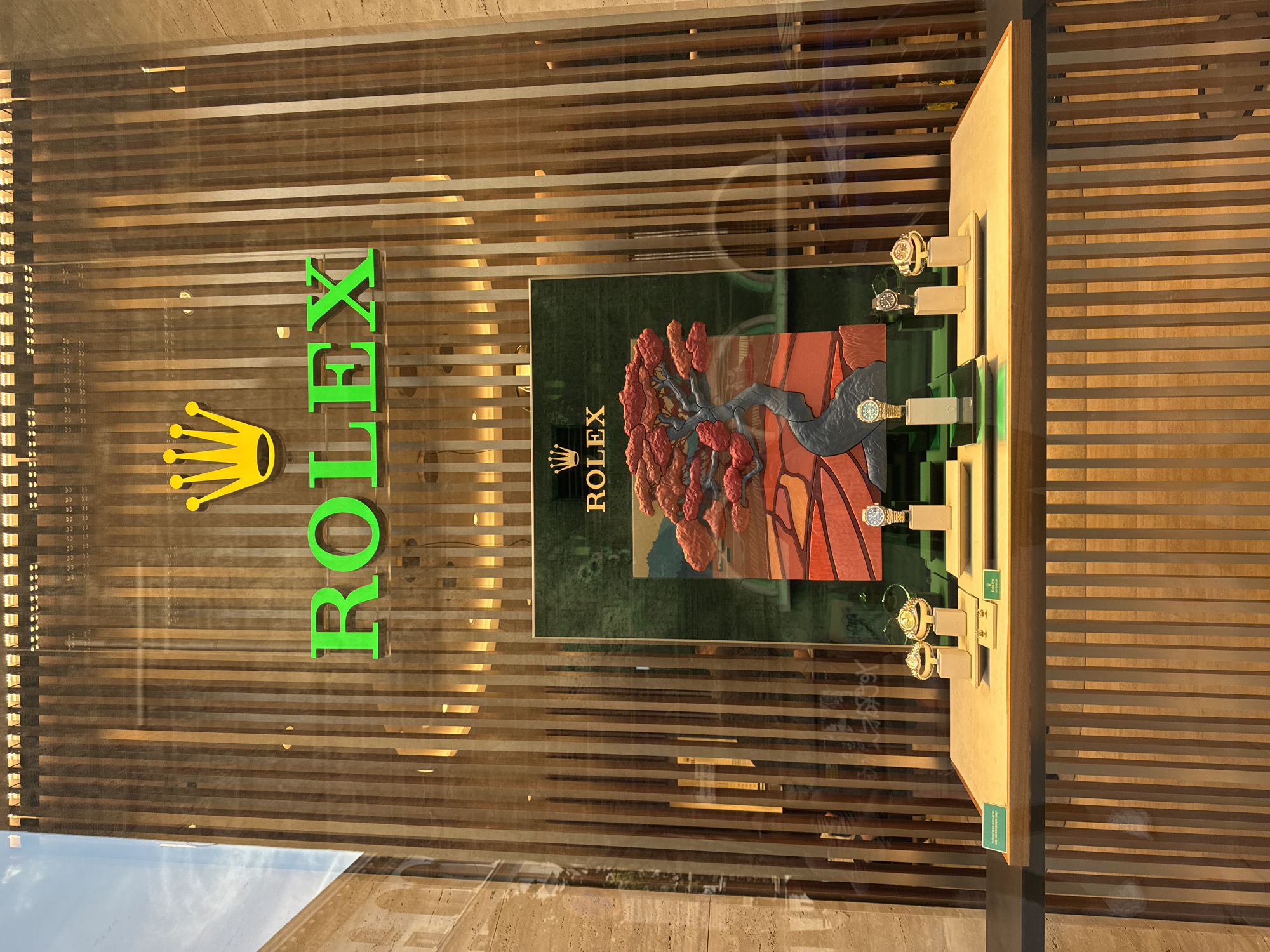 The image displays a Rolex watch store window, viewed from the outside during the daytime. The prominent "ROLEX" logo is affixed to the glass, featuring a yellow crown symbol and bold green lettering. Behind the glass, a textured wooden slatted background is visible, creating a warm, linear pattern.
The main focus of the window display is a large, elaborate art piece positioned centrally. This piece depicts a stylized, ancient-looking tree with vibrant red and orange foliage and a dark, gnarled trunk, set against a dark green backdrop with a reddish-brown, undulating landscape. A smaller white "ROLEX" logo with its crown symbol is visible on the green background of this art piece.
Several Rolex watches are meticulously displayed on individual light-colored stands arranged along the right side of the art piece. From top to bottom, at least four watches are discernible: one with a gold-tone case and white dial, another with a silver-tone case and white dial, followed by a gold-tone watch with a green dial, and another gold-tone watch featuring a fluted bezel. Small green labels with the "ROLEX" brand name are visible on some of the display stands.
Reflections on the shop window show elements of the surrounding environment, including a bright blue sky with white clouds at the bottom left and the beige, textured facade of an adjacent building. The overall impression is one of luxury and careful presentation, with natural light illuminating the display.