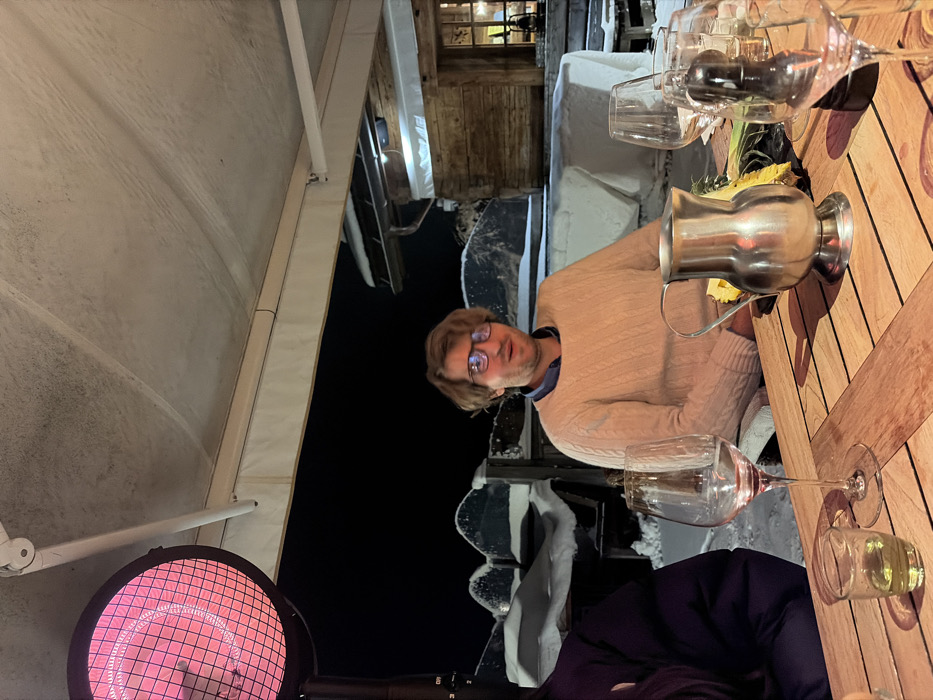 The image depicts an outdoor dining scene at night in La Clusaz, France. The foreground shows a wooden slatted table with several items: an empty wine glass to the left, a small clear shot glass containing a yellow liquid, a silver-colored metal pitcher, and another wine glass along with a black pepper mill on the right. A male individual with light brown hair and eyeglasses, wearing a light beige cable-knit sweater over a collared shirt, is seated at the table, looking forward with a slightly open mouth. To his left, the shoulder and arm of another person, clad in a dark purple or black puffer jacket, are partially visible. A round, red-glowing electric patio heater stands prominently on the far left. Above the scene, a light-colored retractable awning or canopy covers the dining area. The background reveals a snow-covered landscape under a dark night sky. Snow-laden structures, possibly benches or low walls, are visible behind the man. To the far right in the background, a rustic wooden chalet-style building with a lit window can be seen. The overall environment suggests a cold, winter setting, with artificial heating provided for comfort.