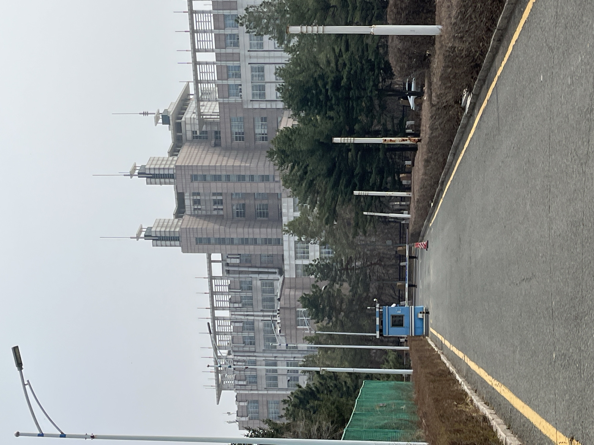 The image depicts a scene in Changchun City, China, featuring a multi-story building, likely an office complex, on the left side. The building has a light brown or grey facade with numerous windows and a tiered structure at the top, with antennas protruding from the top levels. A row of trees partially obscures the view of the building. To the right is an asphalt road with a yellow line marking the edge. A small blue guardhouse stands on the shoulder of the road. White poles, likely lamp posts, are positioned along the side of the road and in front of the building. The sky is overcast, suggesting a cloudy day. The foreground includes a street light and construction netting.