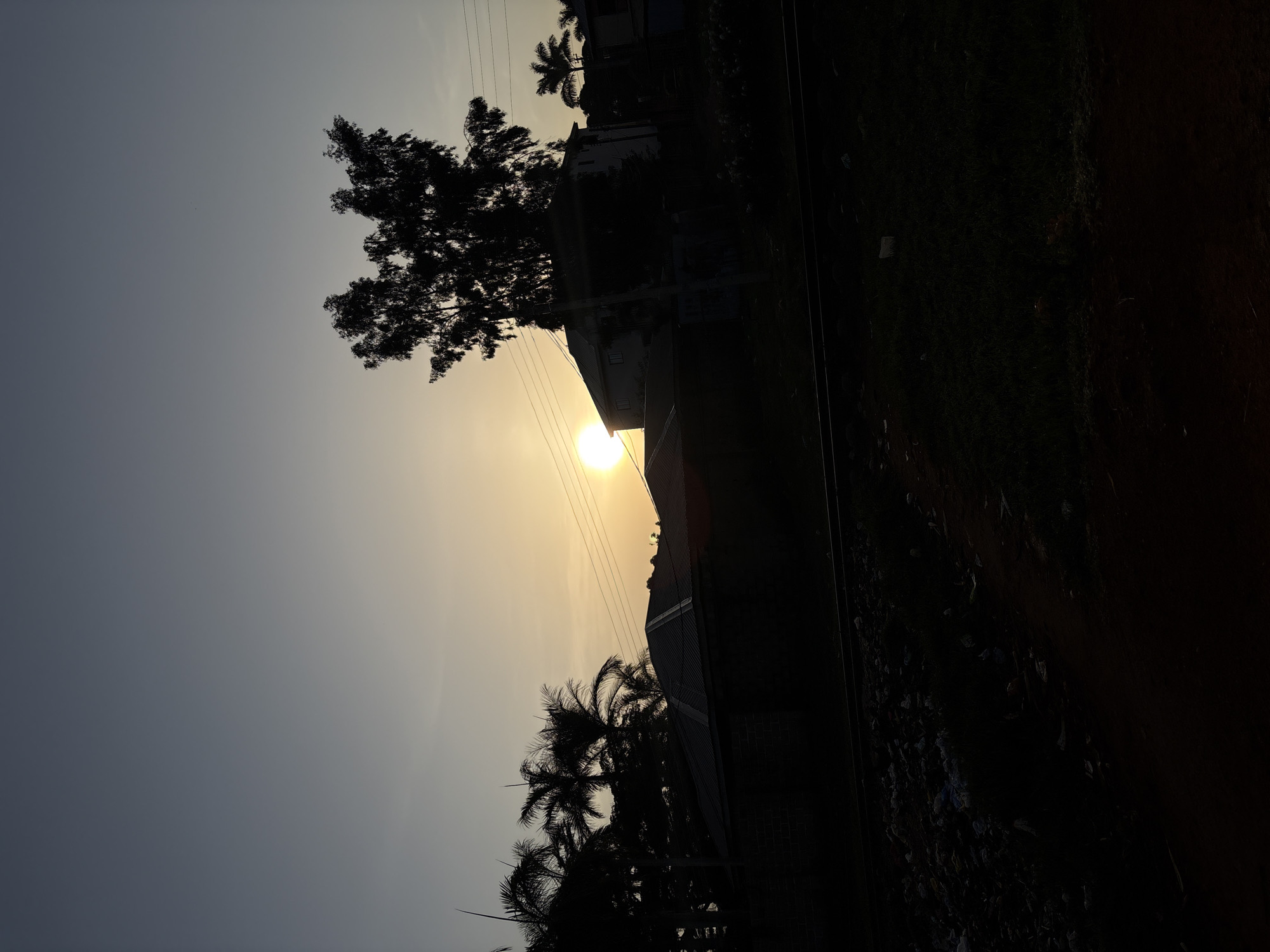 The image depicts a sunset over Bukuru, Nigeria. Silhouetted against the vibrant sky are several buildings, including houses with corrugated iron roofs, and a cluster of palm trees. A large tree with a dense canopy stands prominently in the foreground. Power lines crisscross the scene, adding a utilitarian element to the natural landscape. The ground in the foreground is dark and appears to be a mixture of dirt and debris, possibly indicating an unpaved area or a construction site. The overall atmosphere is tranquil, with the setting sun casting a warm glow over the horizon.