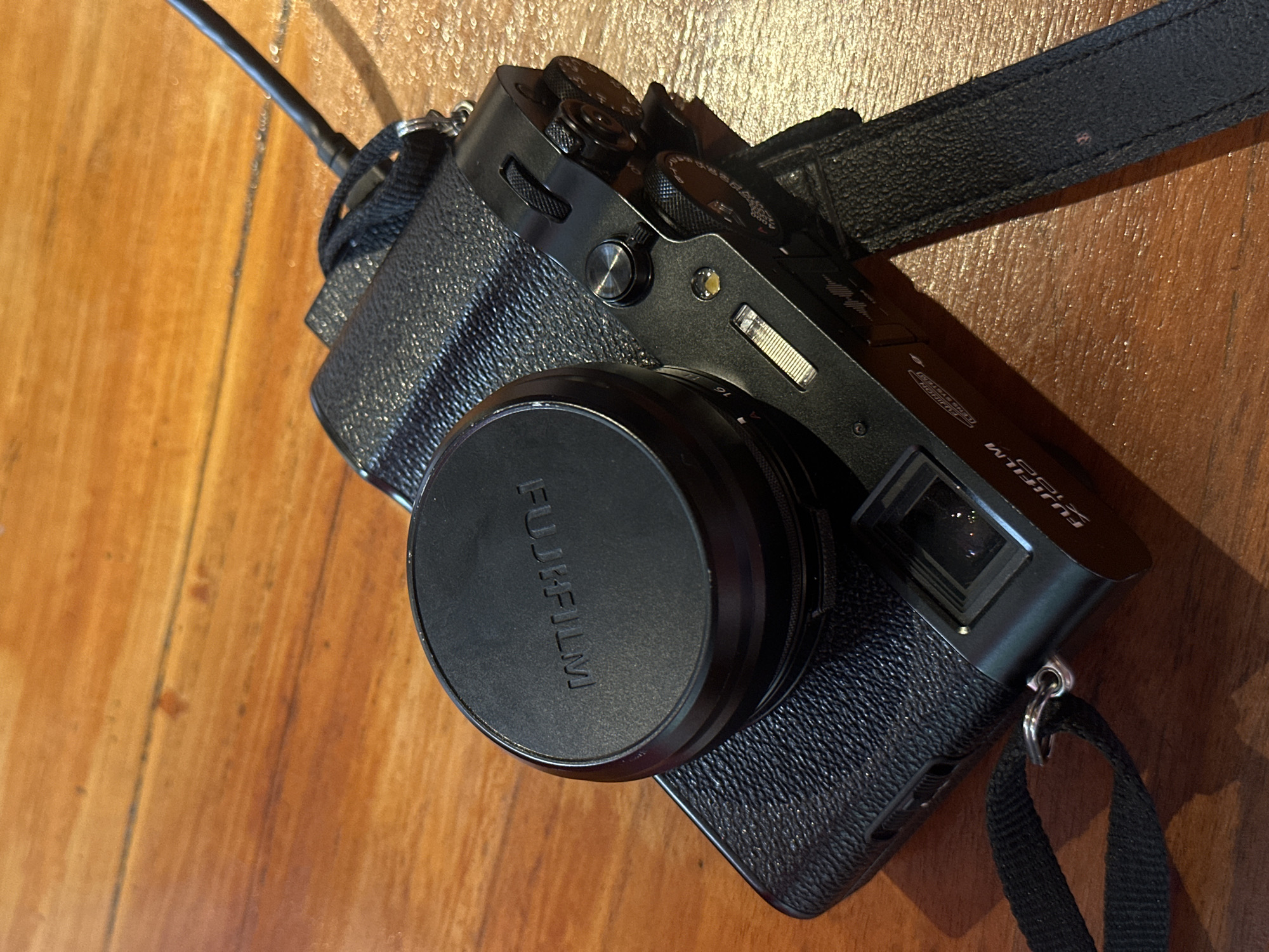 A black Fujifilm X100V camera with a lens cap on is placed on a wooden table. The camera has a textured grip and a black strap attached. The lens cap has the "FUJIFILM" logo embossed on it. The table has a visible wood grain pattern and appears to be a warm, reddish-brown color. The lighting suggests an indoor setting with a light source from the right, casting shadows. The scene is static, with no people or activity visible. The location is Unknown, Unknown.