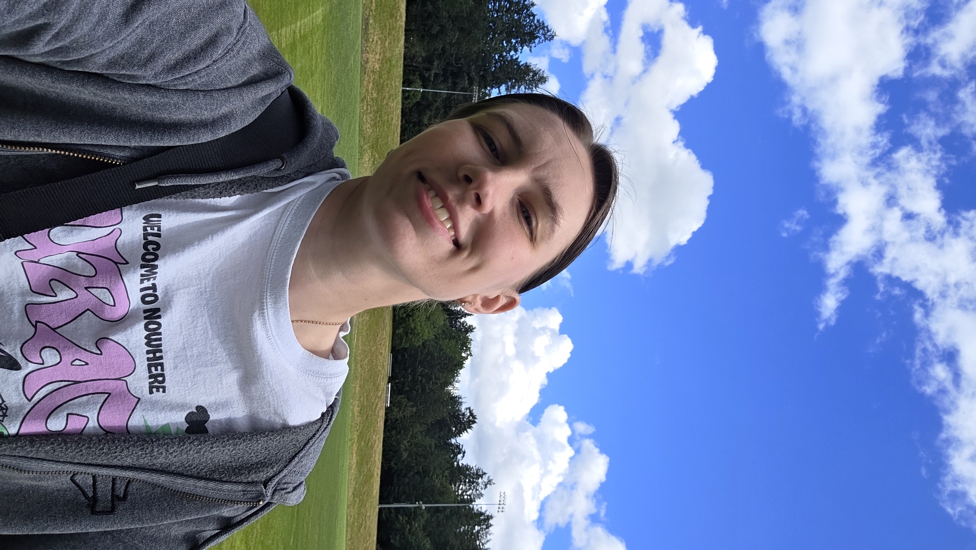 This media file is a selfie of a young person with short brown hair and fair skin, taken outdoors on a bright, sunny day. They are smiling and looking directly at the camera. The person is wearing a grey zip-up hoodie over a white t-shirt. The t-shirt has a graphic design with large, stylized pink lettering and the text "WELCOME TO NOWHERE" printed vertically in black.
The background features a grassy field, likely a park or sports field, with a line of dark green trees in the distance. The sky above is a vibrant blue, dotted with fluffy white clouds. There are some stadium lights visible behind the trees, suggesting a sports venue. The overall impression is one of a casual, outdoor activity, possibly a break or a moment of relaxation during a sunny day in Portland, USA. The bright lighting and blue sky indicate it is daytime, likely late morning or early afternoon. The person appears happy and relaxed.